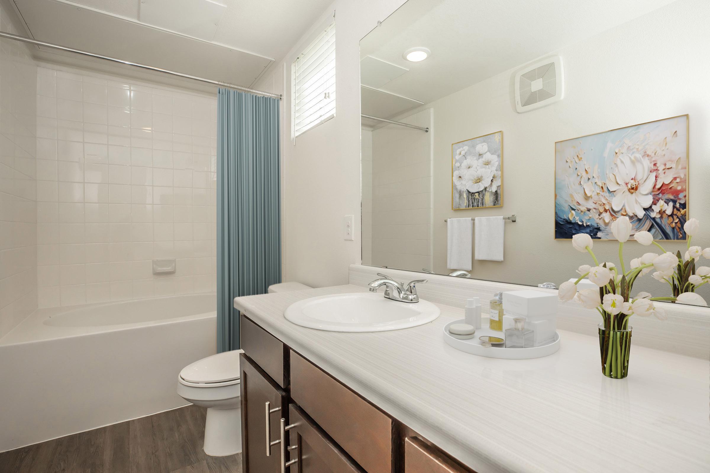 A neatly organized bathroom featuring a white bathtub with a blue shower curtain, a white countertop with toiletries, and a mirror. Decorative flowers are placed on the counter, and a floral artwork decorates the wall. Light streams in from a window, creating a bright and clean atmosphere.