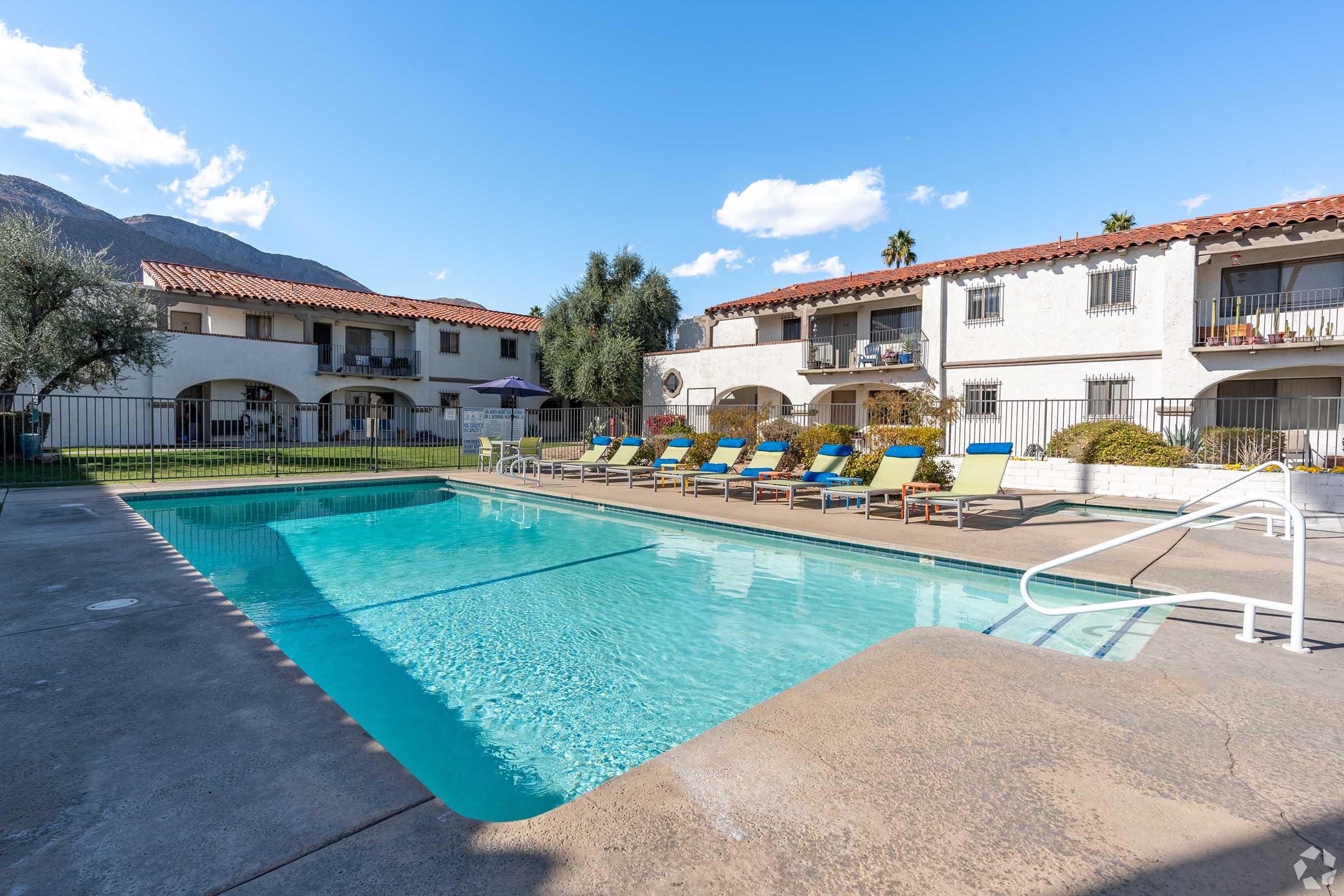 A serene swimming pool surrounded by lounge chairs, with two-story white buildings in the background. The scene features palm trees and mountains under a clear blue sky, creating a relaxing atmosphere. The area is well-maintained and inviting, perfect for leisure and relaxation.