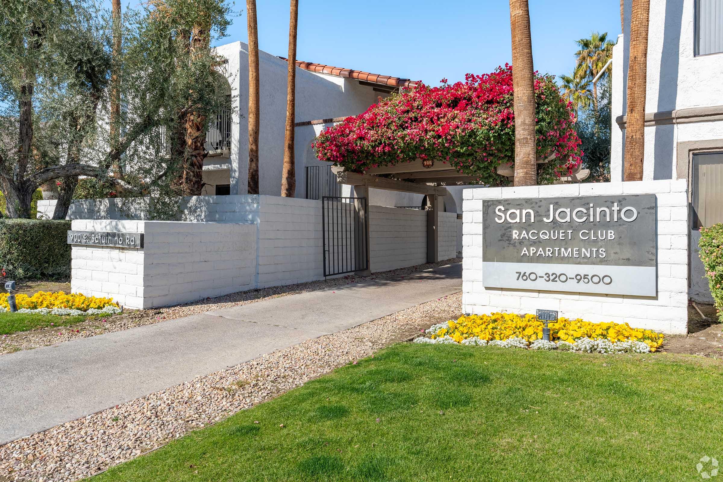 Entrance to San Jacinto Racquet Club Apartments featuring a sign with the property name and contact number. Lush landscaping with vibrant flowers lines the pathway, and palm trees add a tropical feel, creating a welcoming atmosphere.