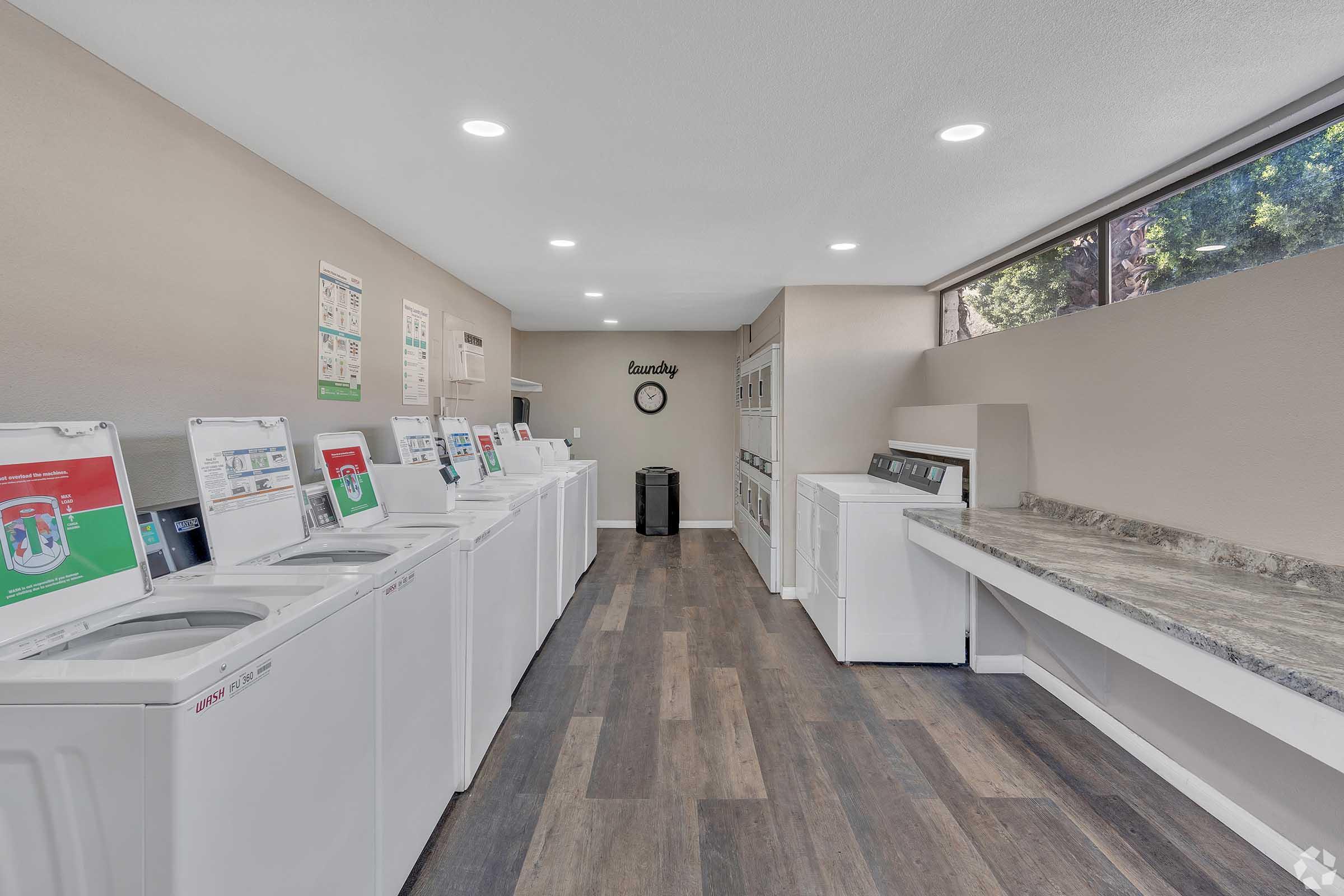 A clean and well-lit laundry room featuring multiple white washing machines and dryers along one wall. There's a countertop for folding clothes and a clock on the wall. Large windows allow natural light to brighten the space, creating an inviting atmosphere. Laundry-related posters are displayed on the walls.