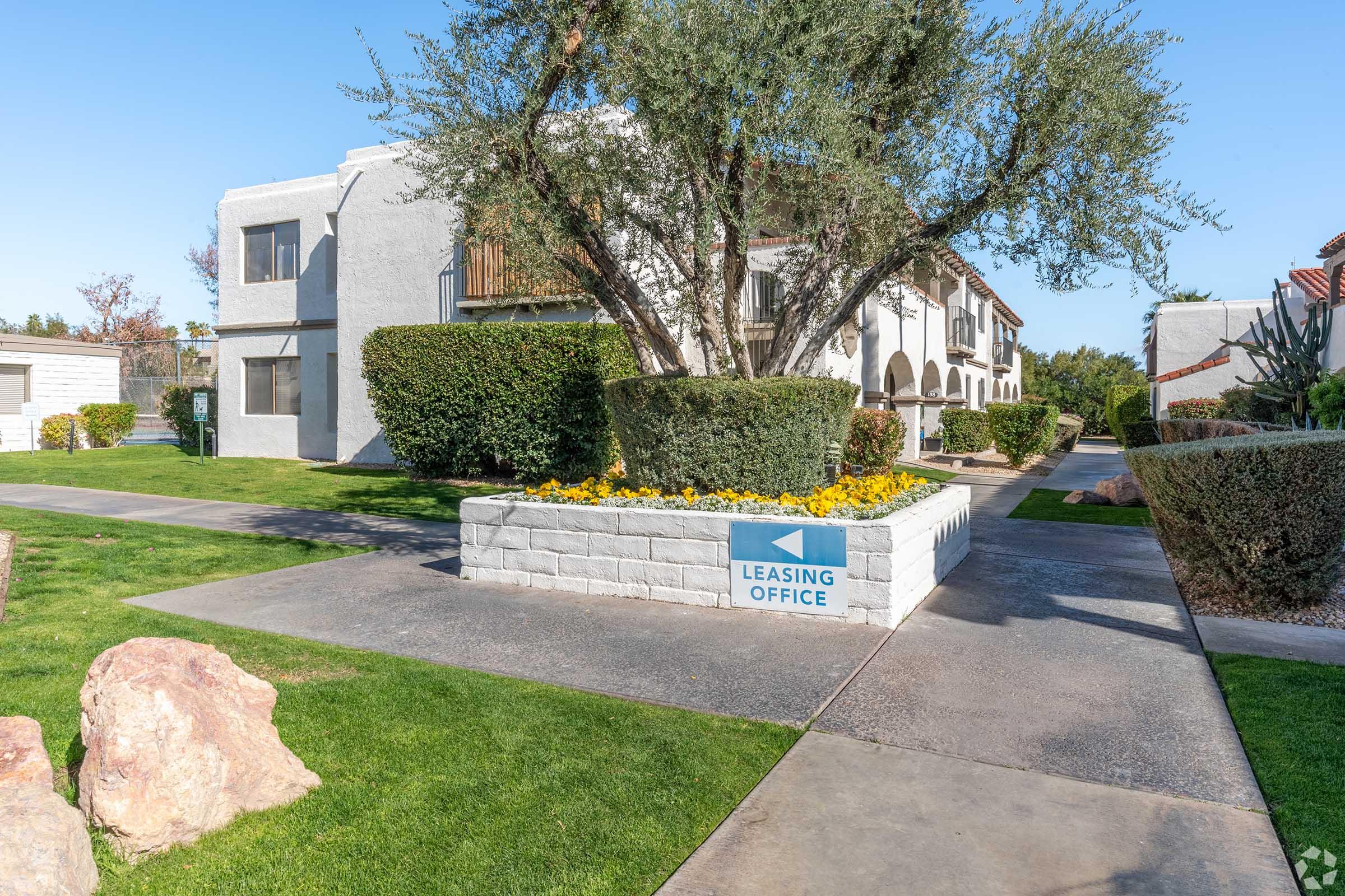 A well-maintained apartment complex featuring a leasing office with a blue sign pointing to it. The area is landscaped with green grass, flowering plants, and decorative rocks, surrounded by residential buildings. Sunlight casts a warm glow on the pathway leading to the office.