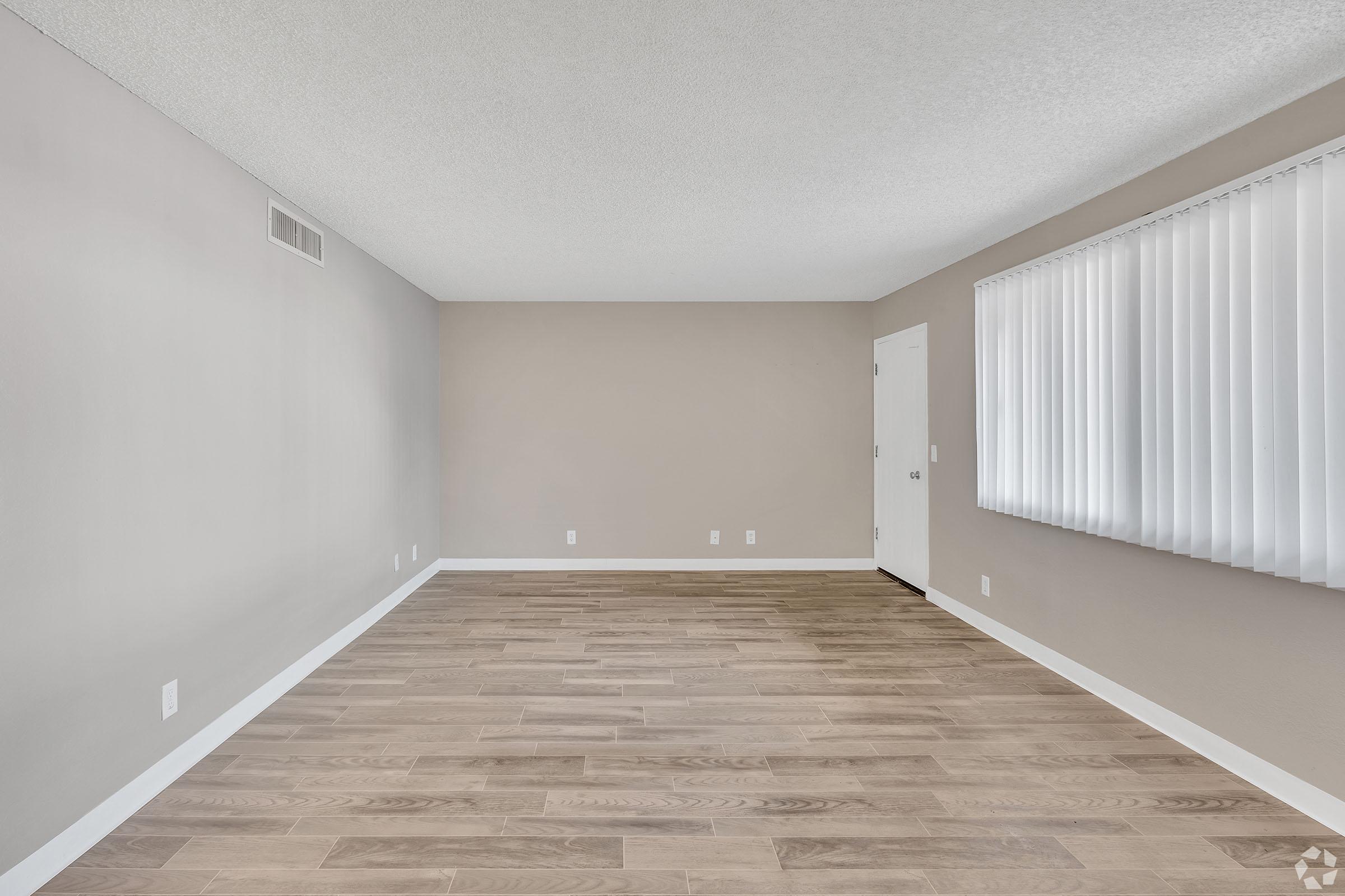Empty room with light gray walls and a smooth wooden floor. A large window with vertical blinds allows natural light to enter. The left side shows a door and the right side features a baseboard. The overall space is uncluttered, offering a modern and minimalistic look.