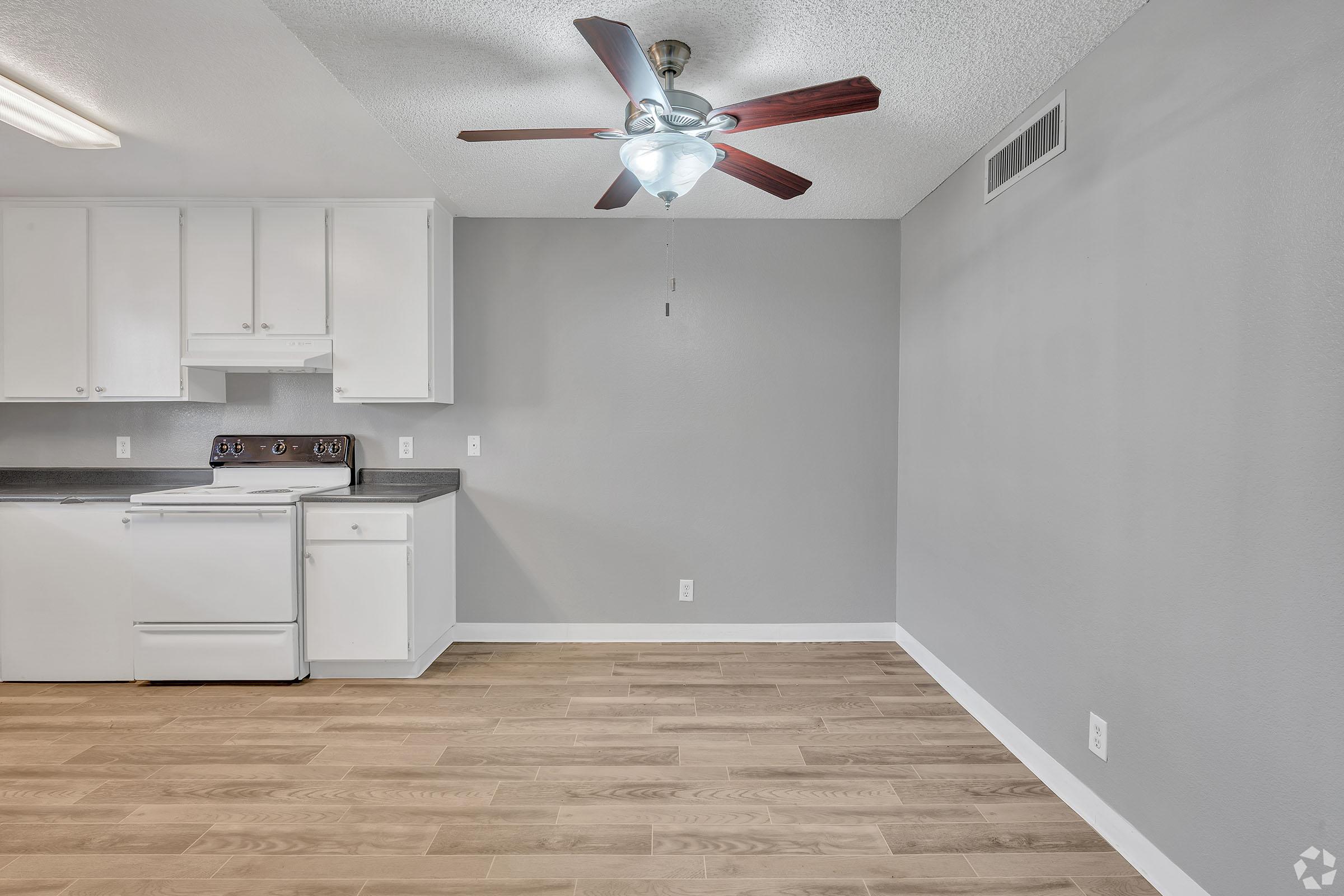 Interior view of a modern kitchen and living area with light-colored wood flooring. The kitchen features white cabinets, a stove, and a refrigerator. A ceiling fan with light is mounted above, and the walls are painted in a soft gray color. The space is bright and open, ideal for a comfortable living environment.
