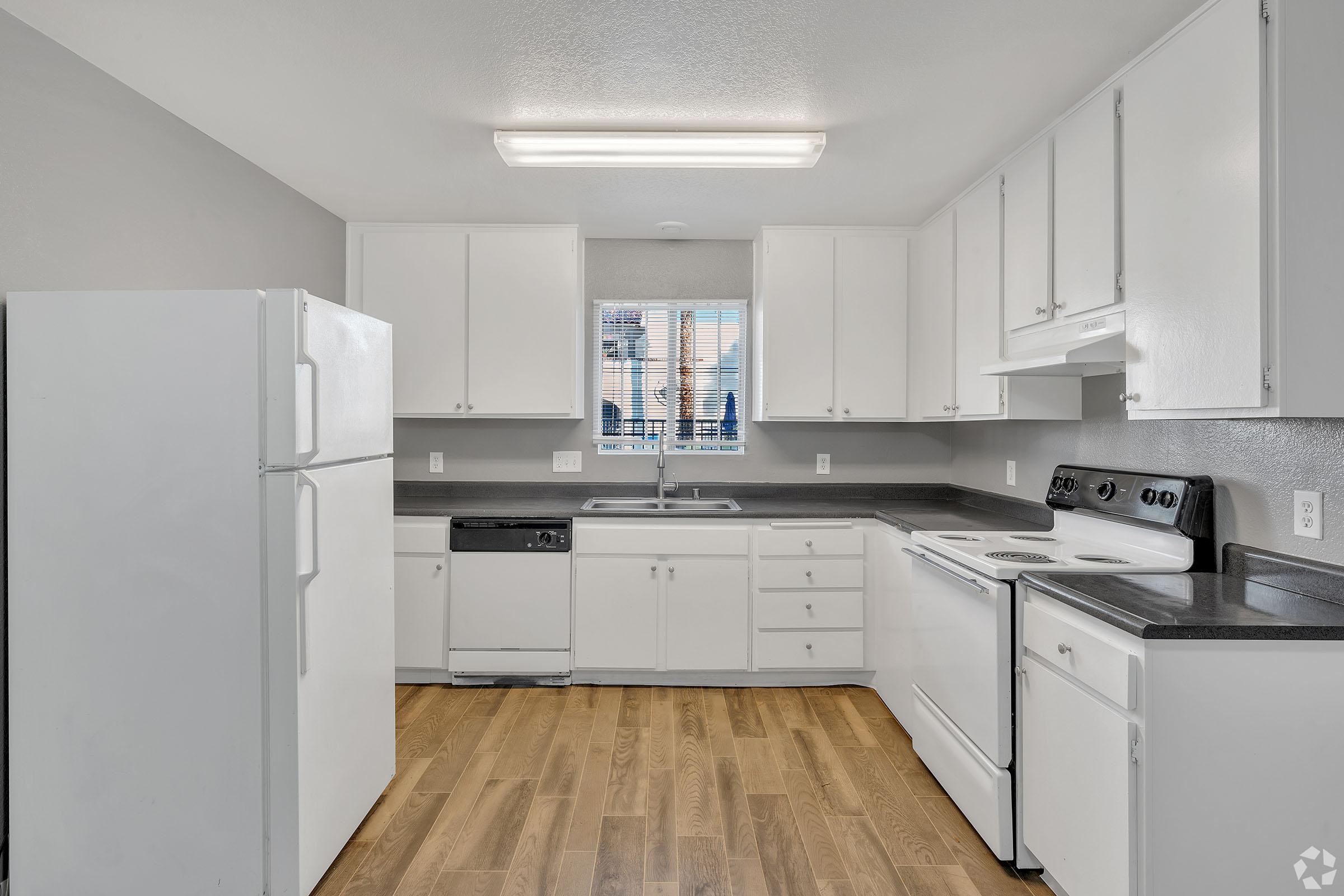 A clean, modern kitchen featuring white cabinetry, a white refrigerator, an oven, and a dishwasher. The countertop is dark, contrasting with the light cabinets. Natural light comes through a window above the sink, and the floor is made of light wood laminate. The space is organized and minimalistic.