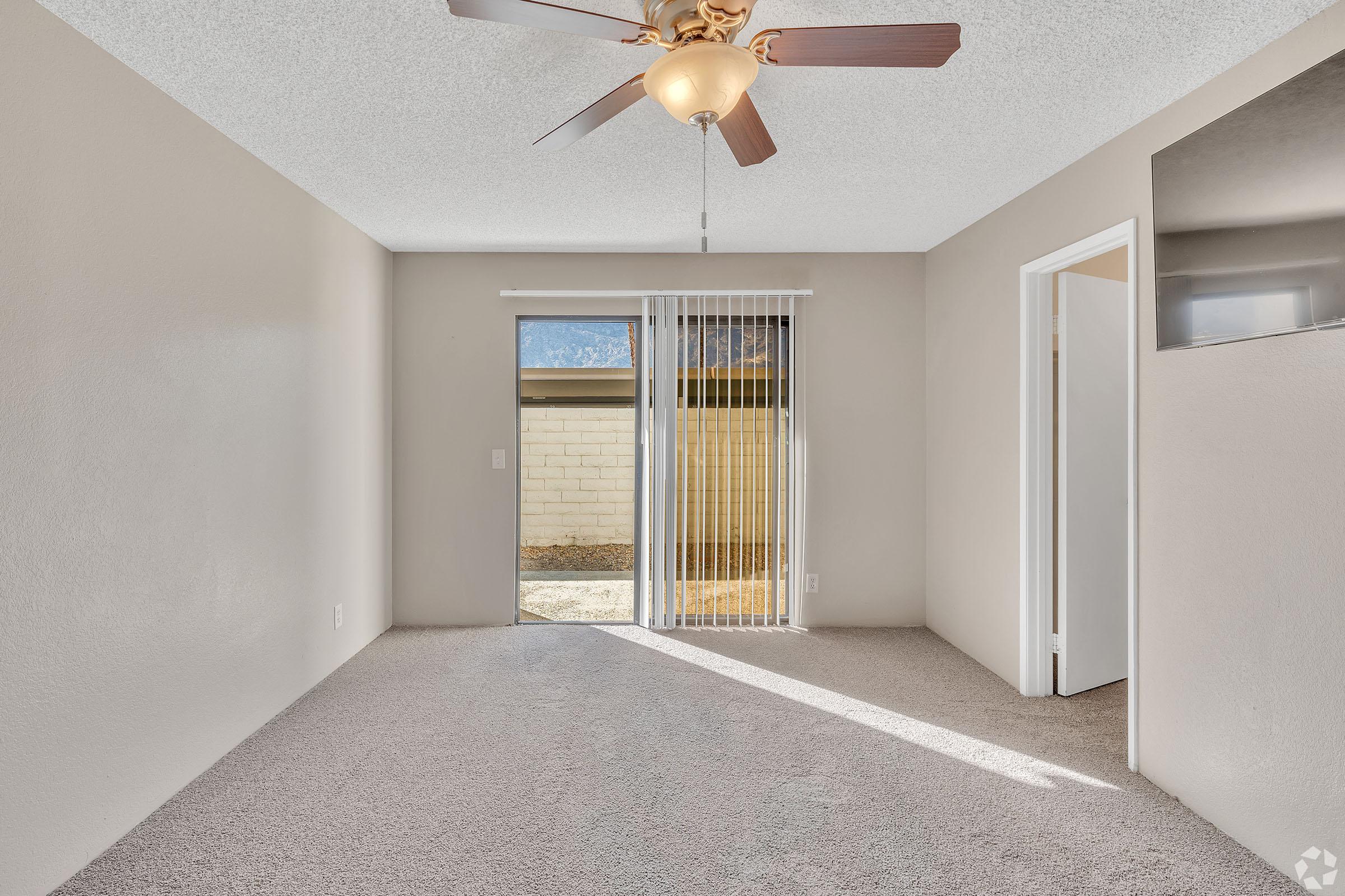 Interior view of a light-colored room with a ceiling fan and carpeted flooring. A sliding glass door leads to an outdoor area, and a doorway is visible on the right. Natural light enters the space, highlighting the neutral-colored walls.