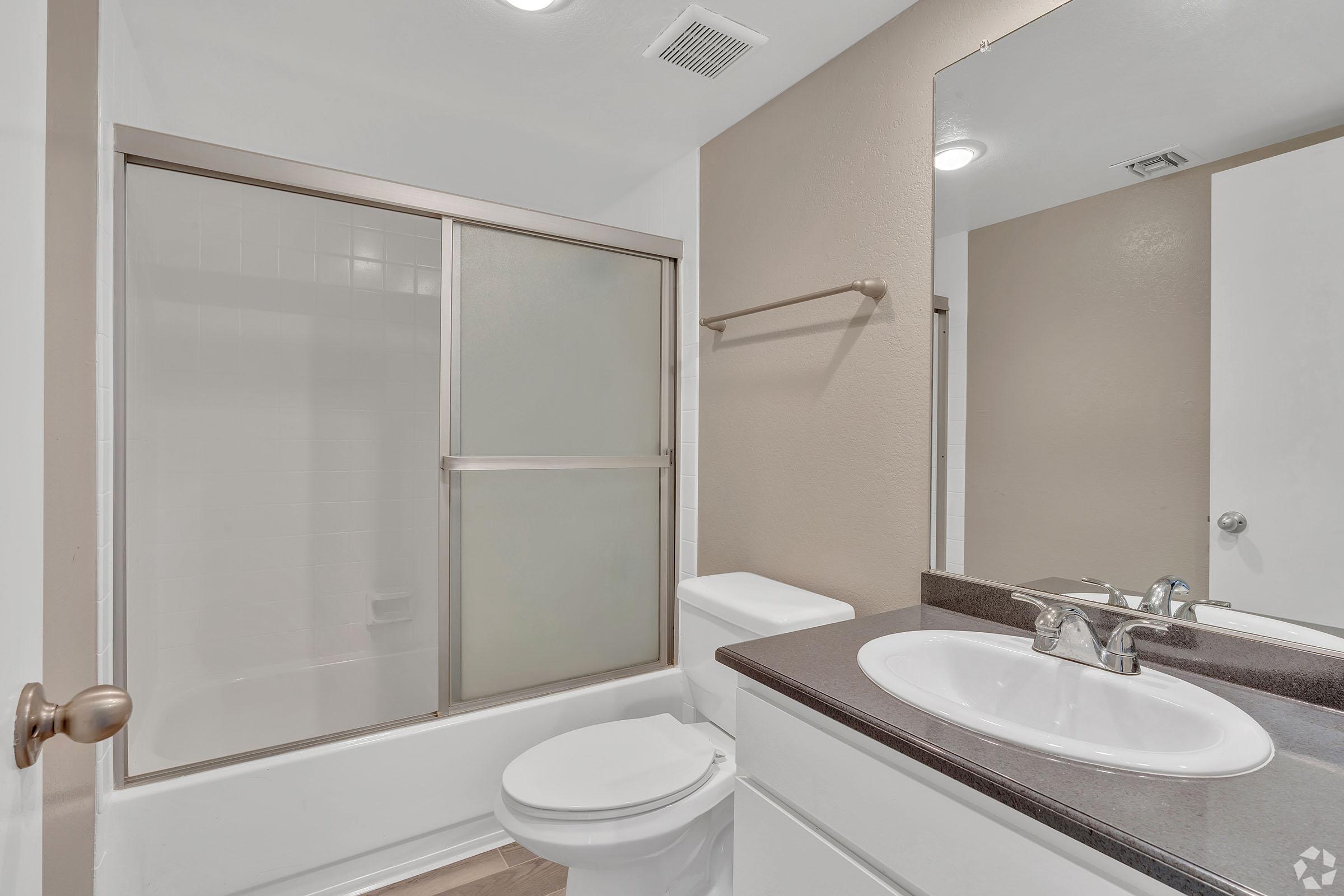 A modern bathroom featuring a glass shower enclosure, a white toilet, and a sleek vanity with a dark countertop and a sink. The walls are painted in a light beige color, creating a clean and contemporary look. A large mirror is mounted above the vanity, reflecting the space.