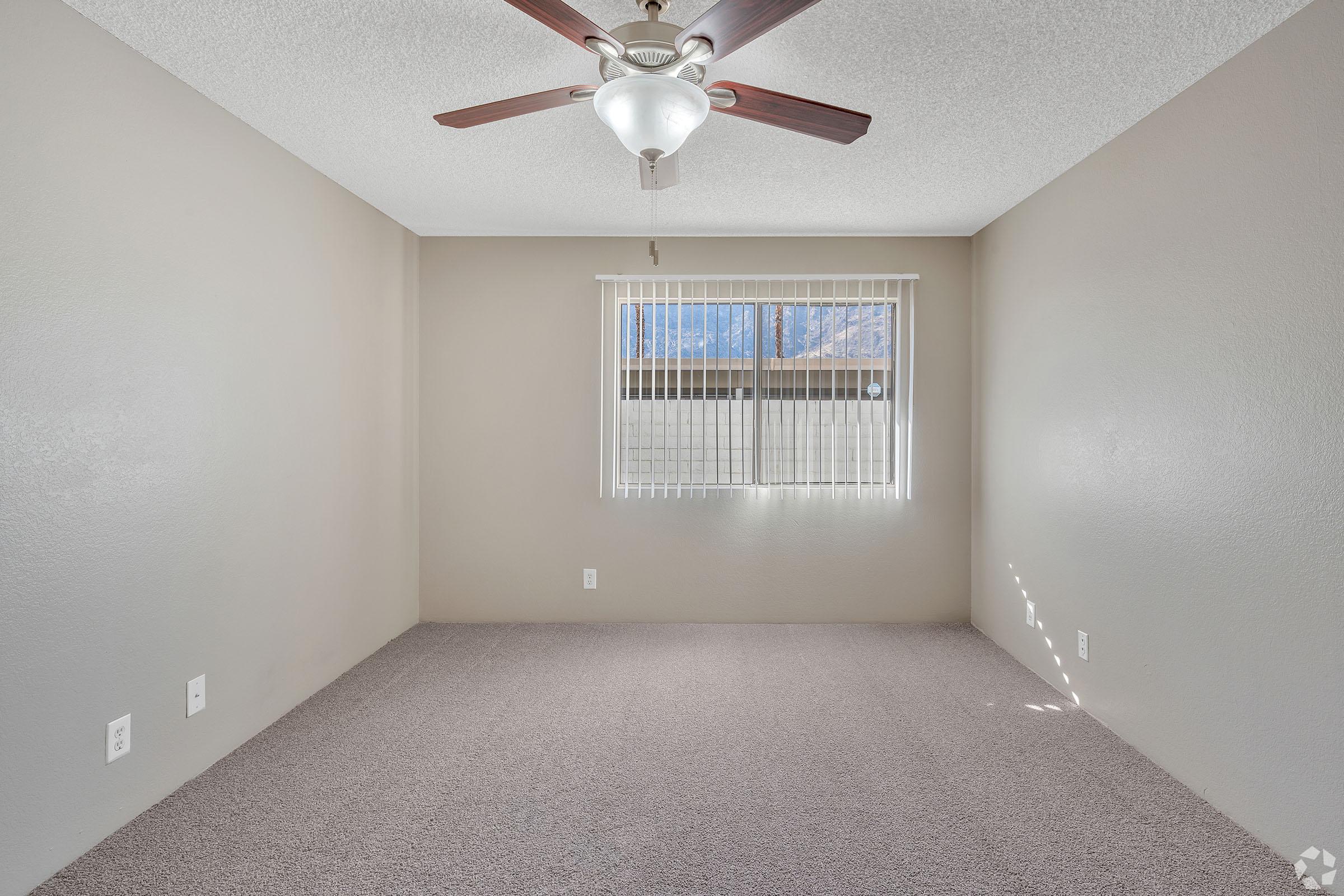 A well-lit, empty room featuring beige walls and a ceiling fan with wooden blades. The floor is covered in light-colored carpet, and there are vertical blinds on a window allowing natural light to enter. The room has a clean and spacious appearance, suitable for various furnishings or uses.