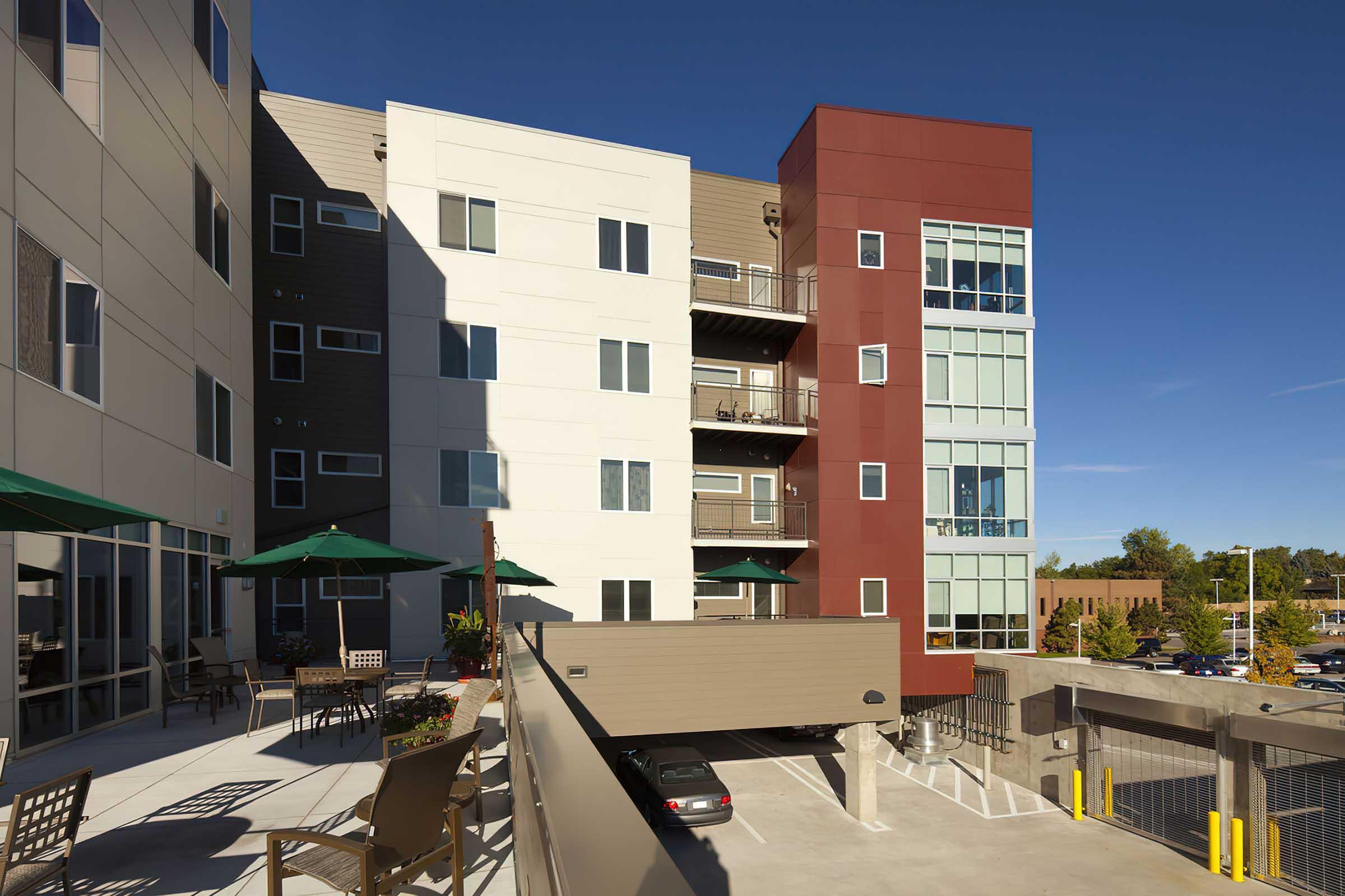 Modern multi-story apartment building featuring a mix of beige and red siding. Balconies extend from several units, and outdoor seating areas with green umbrellas are visible.