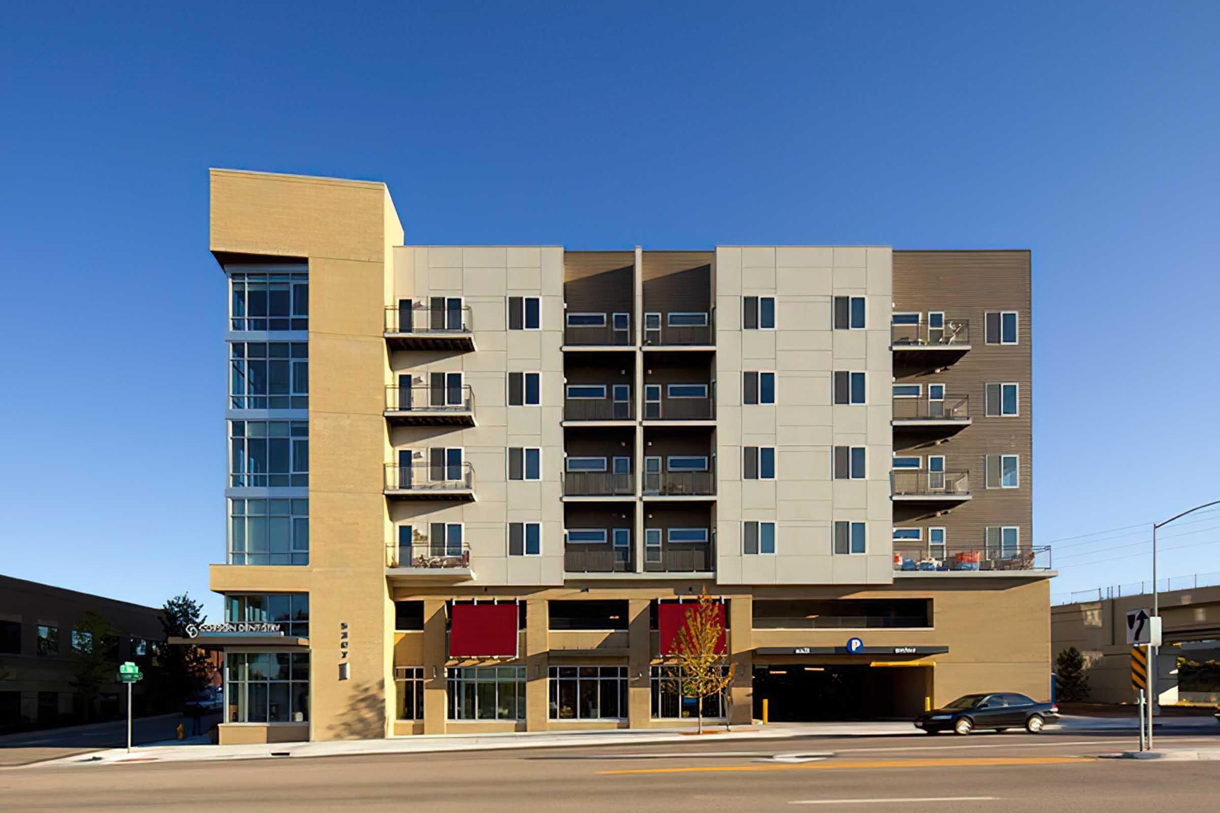 Modern five-story apartment building with a mix of beige and gray panels. Ground level features large glass windows and a restaurant with red awnings.