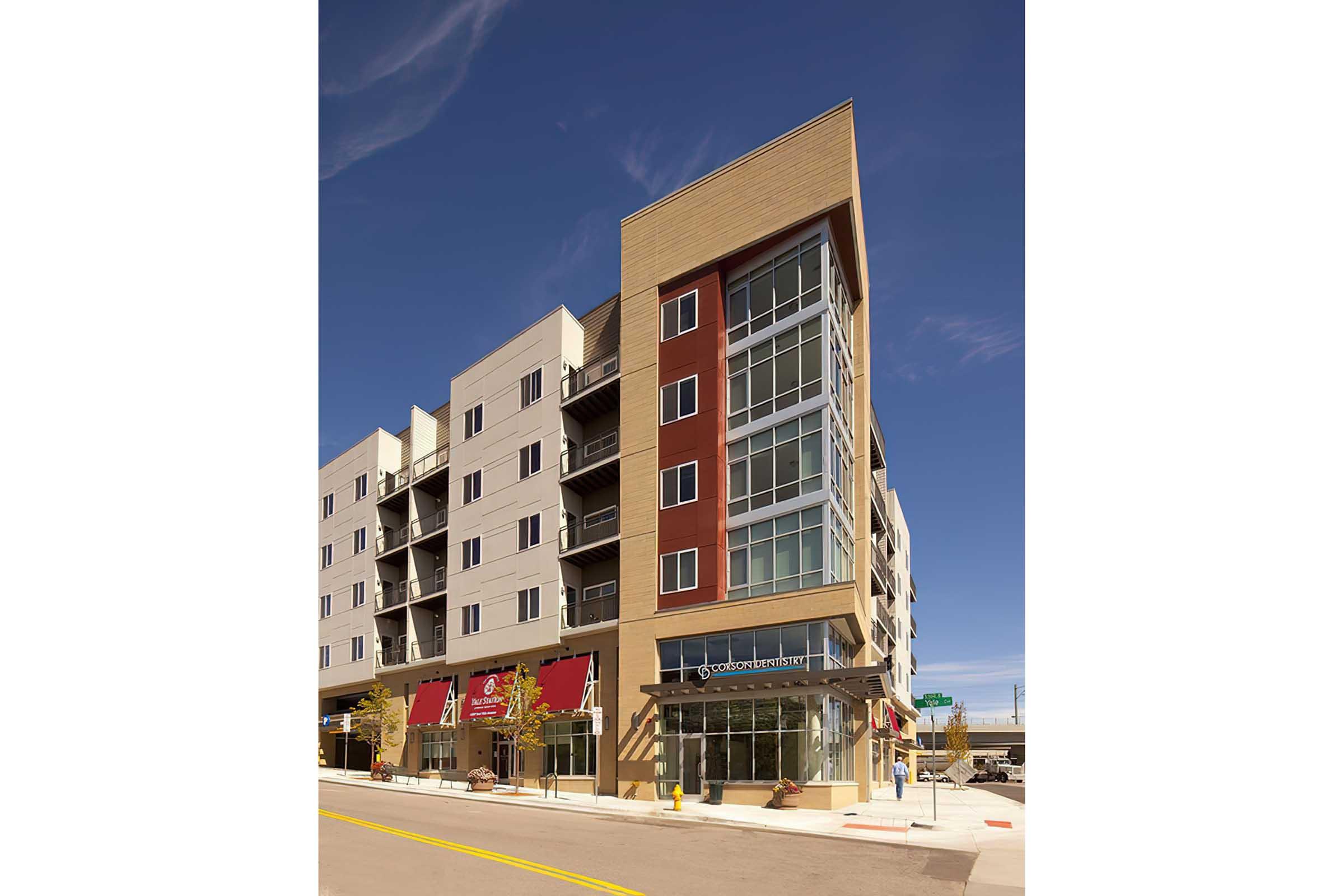 Modern multi-story apartment building with a mix of light and dark siding, featuring large windows and a ground-level retail space.