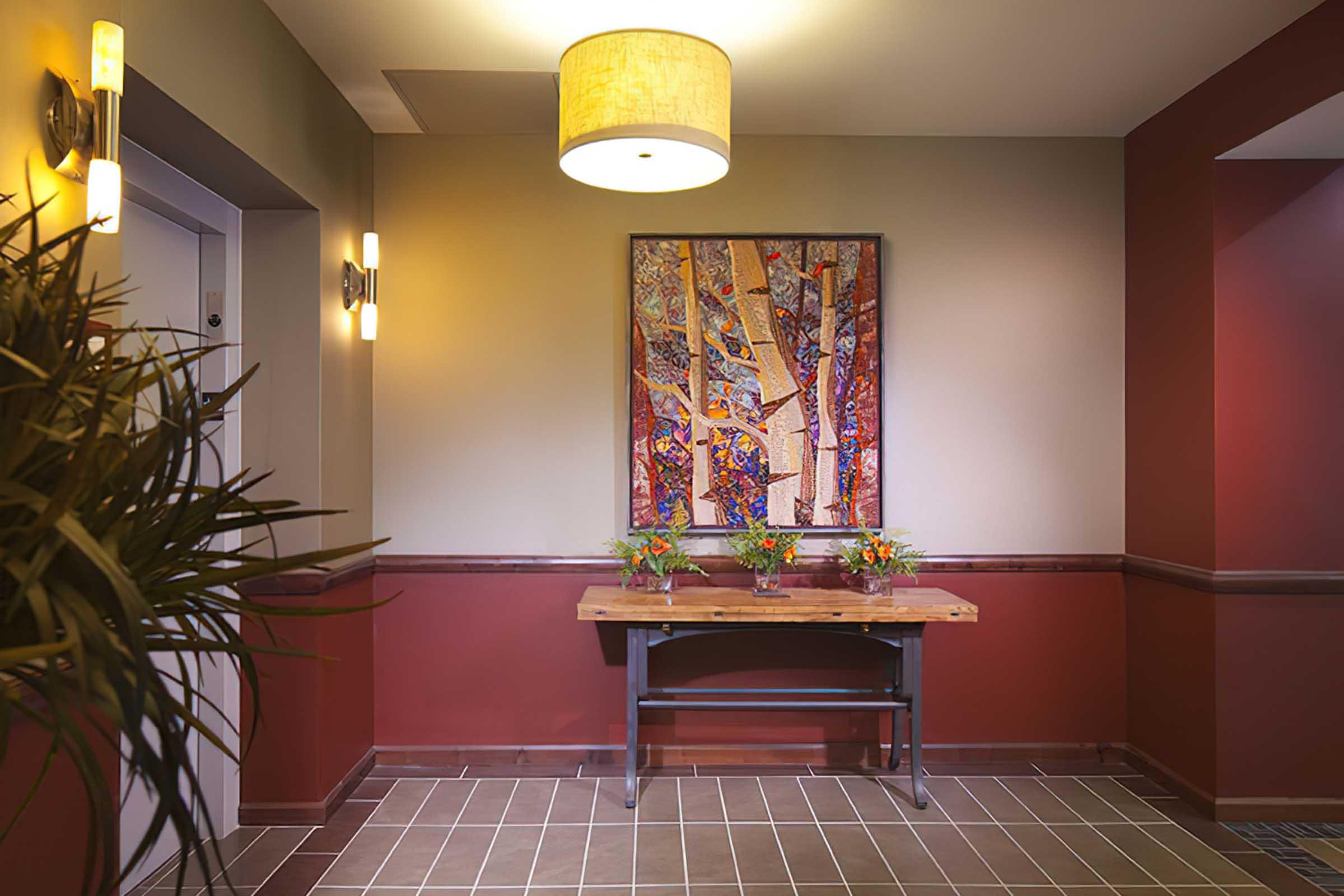 A well-lit hallway featuring an elevator door, a large colorful abstract painting on the wall, and a wooden table with small flower arrangements.