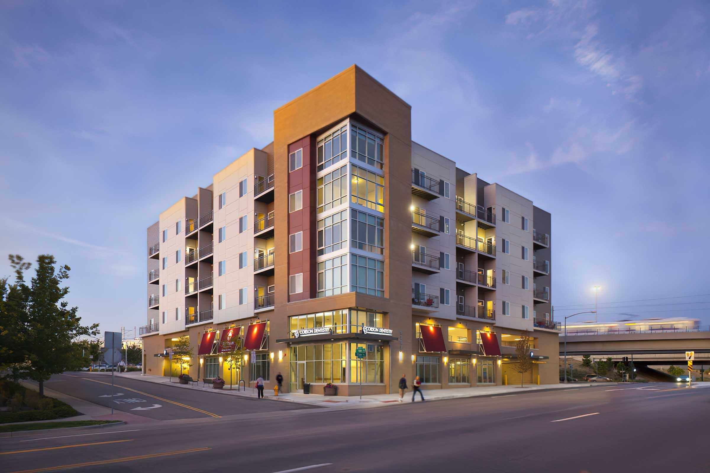 Modern multi-story apartment building at dusk, featuring a mix of brick and light-colored siding. Ground-level retail spaces with large windows and outdoor seating areas are visible.
