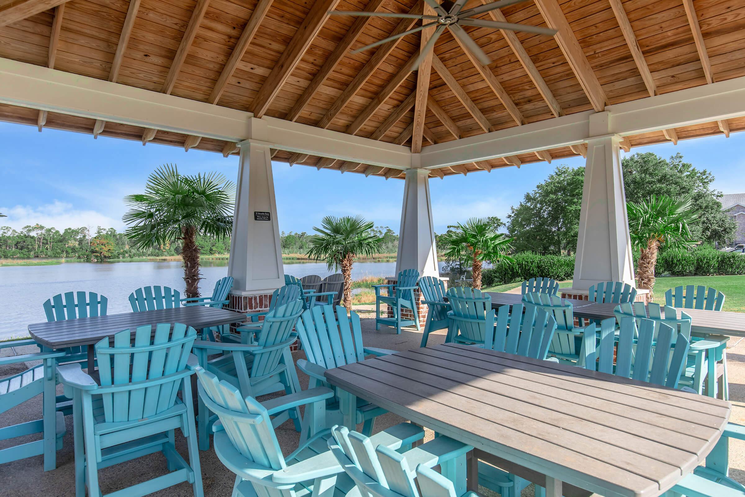 A covered outdoor seating area with turquoise chairs and wooden tables, overlooking a calm lake surrounded by greenery and palm trees. The space features a high wooden ceiling with a fan, providing a relaxed atmosphere for gatherings by the water.