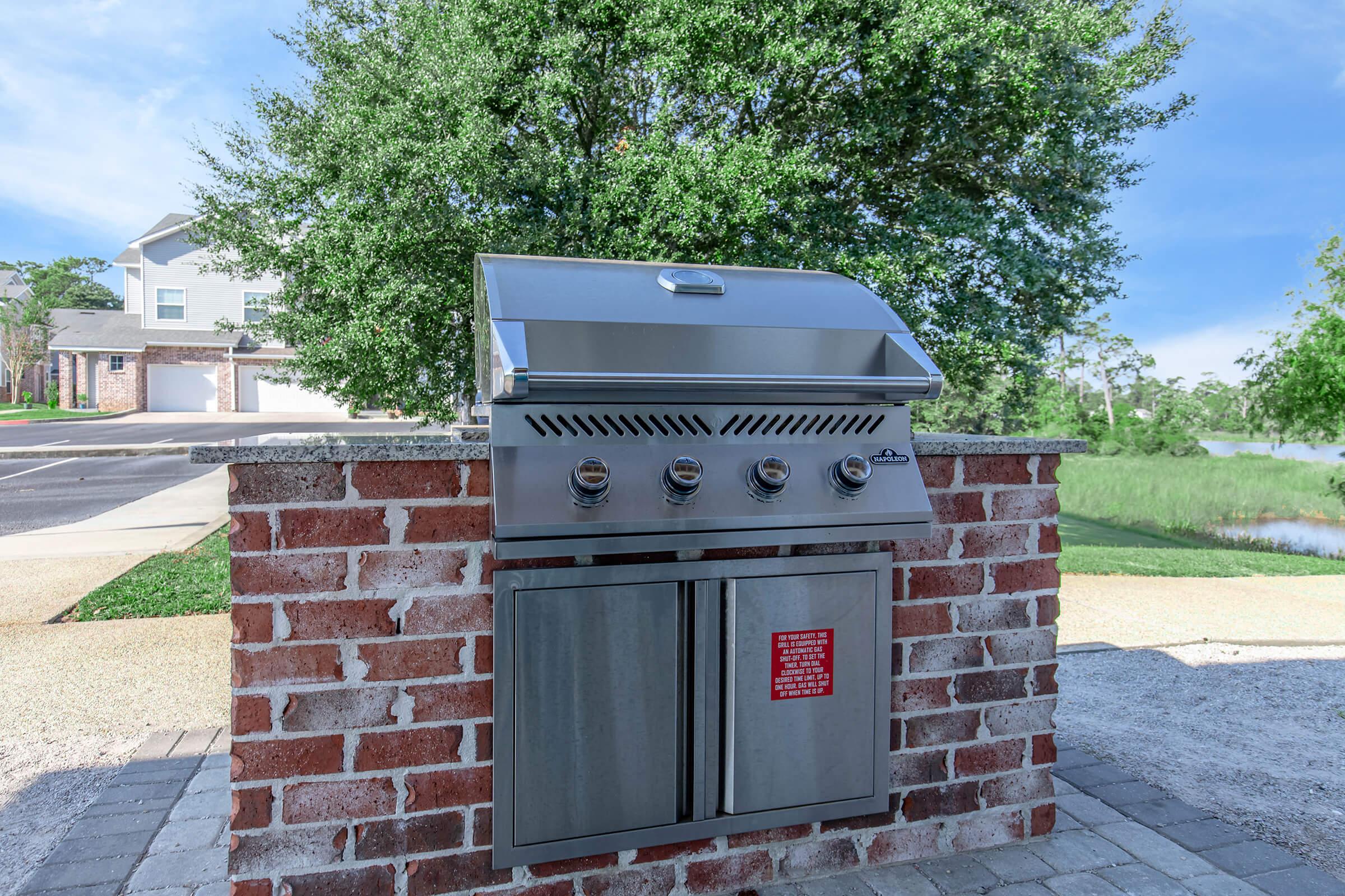 A silver outdoor grill mounted on a brick enclosure, featuring three burners and a closed storage area below. The background shows a scenic view with trees and a body of water, along with a paved area and nearby buildings. The sky is clear and blue.