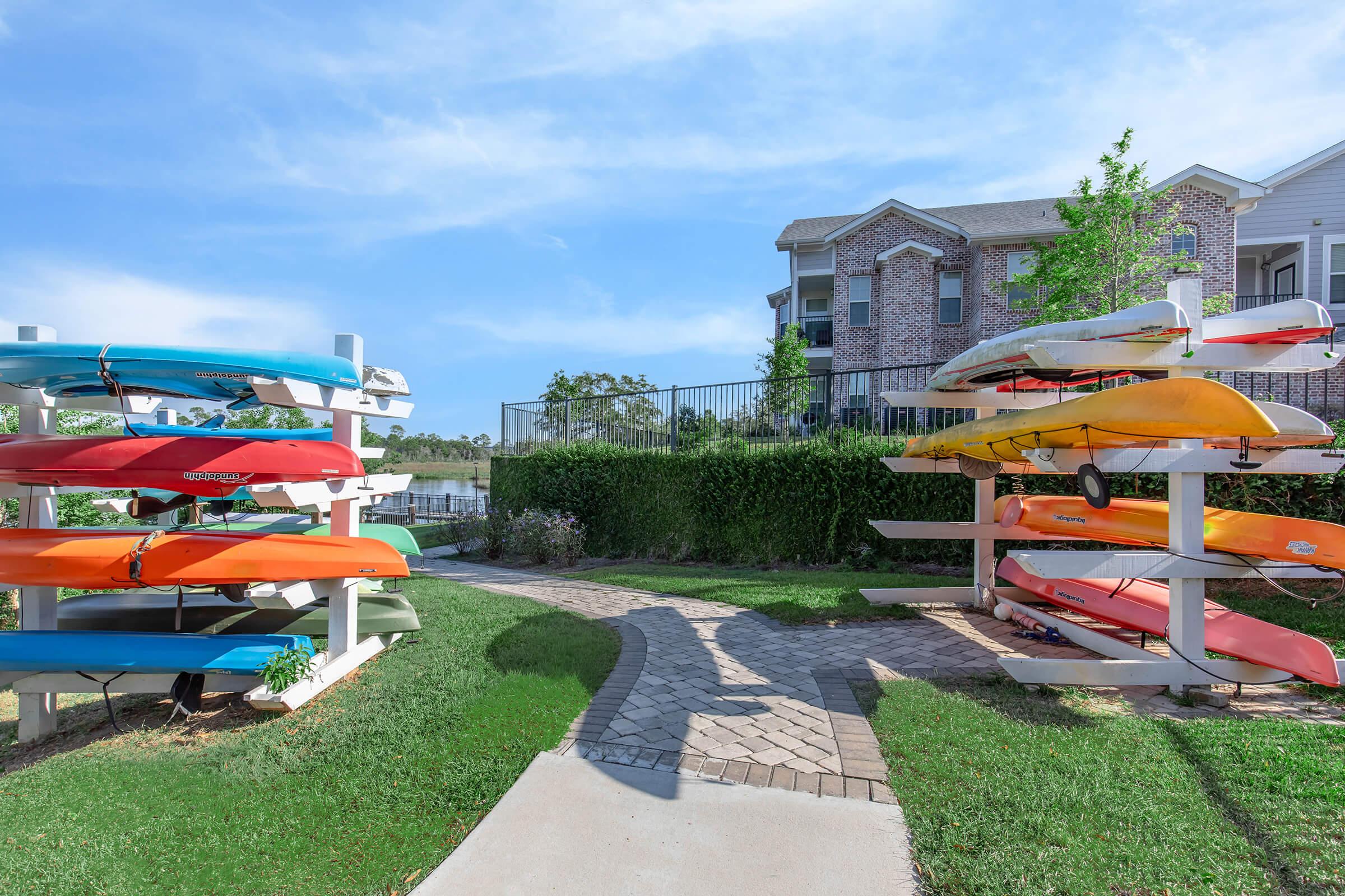 Colorful kayaks and canoes are neatly stacked on racks beside a grassy pathway leading to a waterway. A brick path runs through the scene, with trees and shrubs in the background, and a multi-story building visible beyond the kayaks under a clear blue sky.