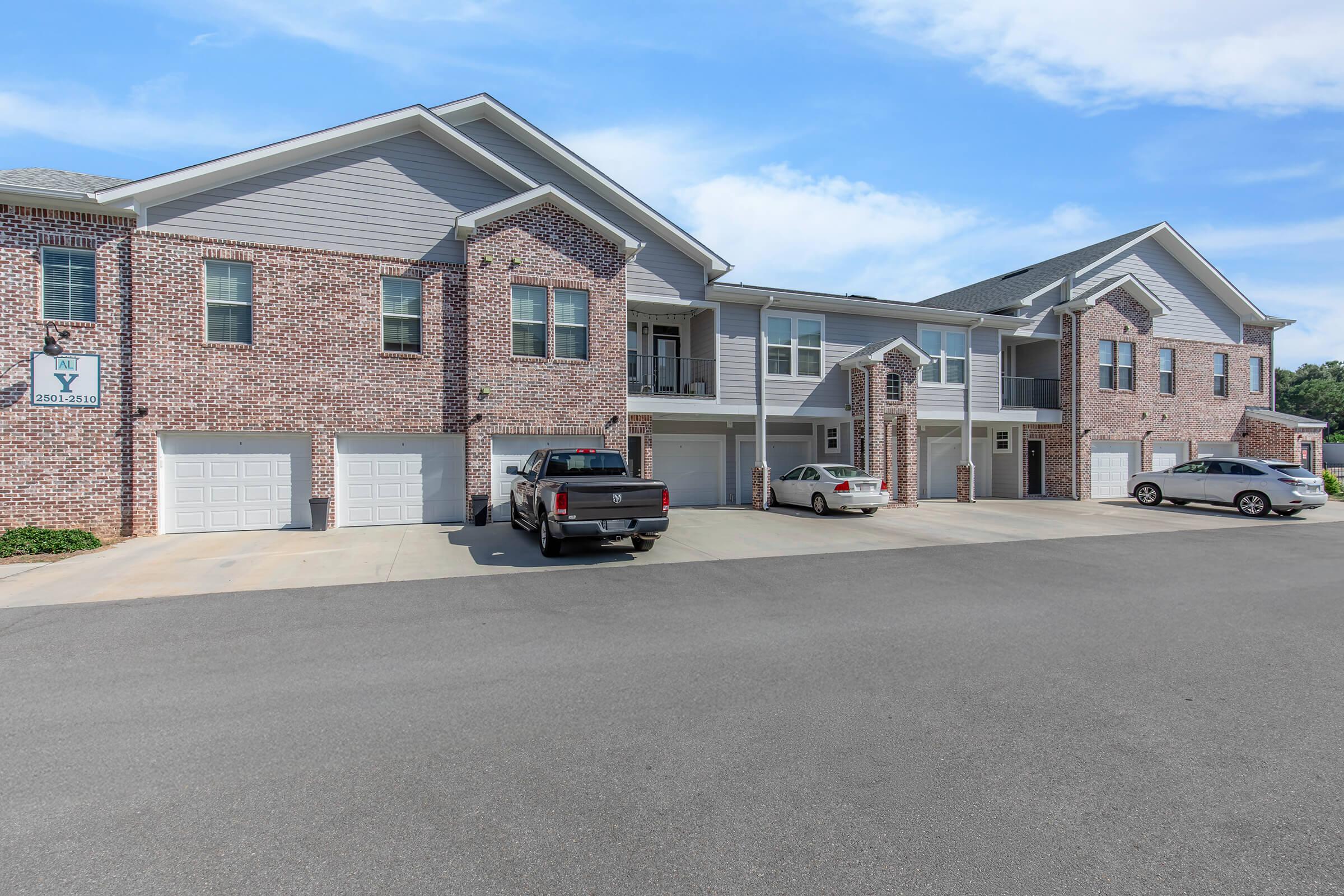 A row of brick apartment buildings with gray accents, featuring multiple balconies and garages. The parking lot has several vehicles, including a black pickup truck and a white car. The sky is clear with a few clouds, and the surroundings appear well-maintained.