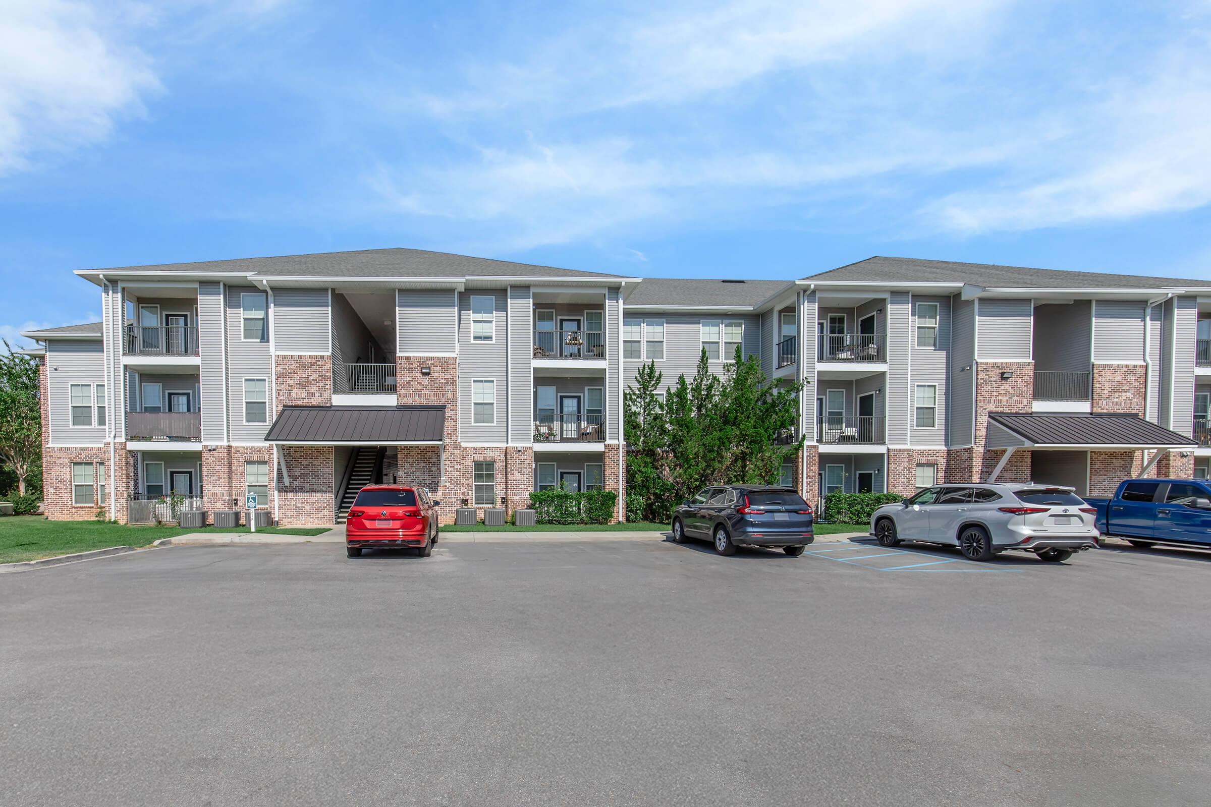 Three-story apartment building with a mix of light gray and brick exterior. Several parked cars are visible in the foreground, including a red SUV. The building features balconies on each floor and landscaped greenery at the base. The sky above is clear with a few clouds.