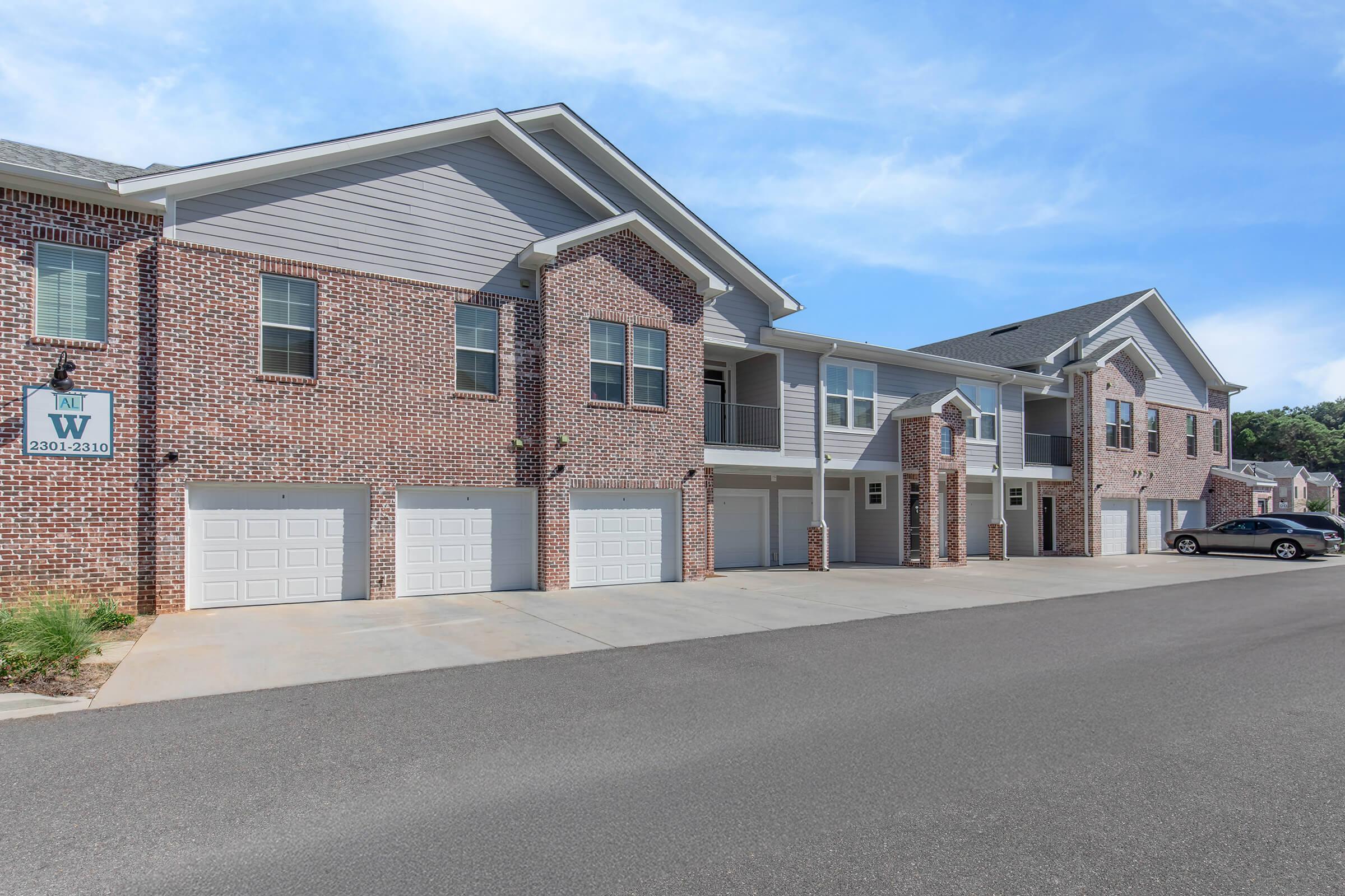 A modern brick apartment building featuring multiple units with balconies, garages on the ground level, and a smooth paved driveway. The sky is clear with a hint of blue, creating a bright and inviting atmosphere.