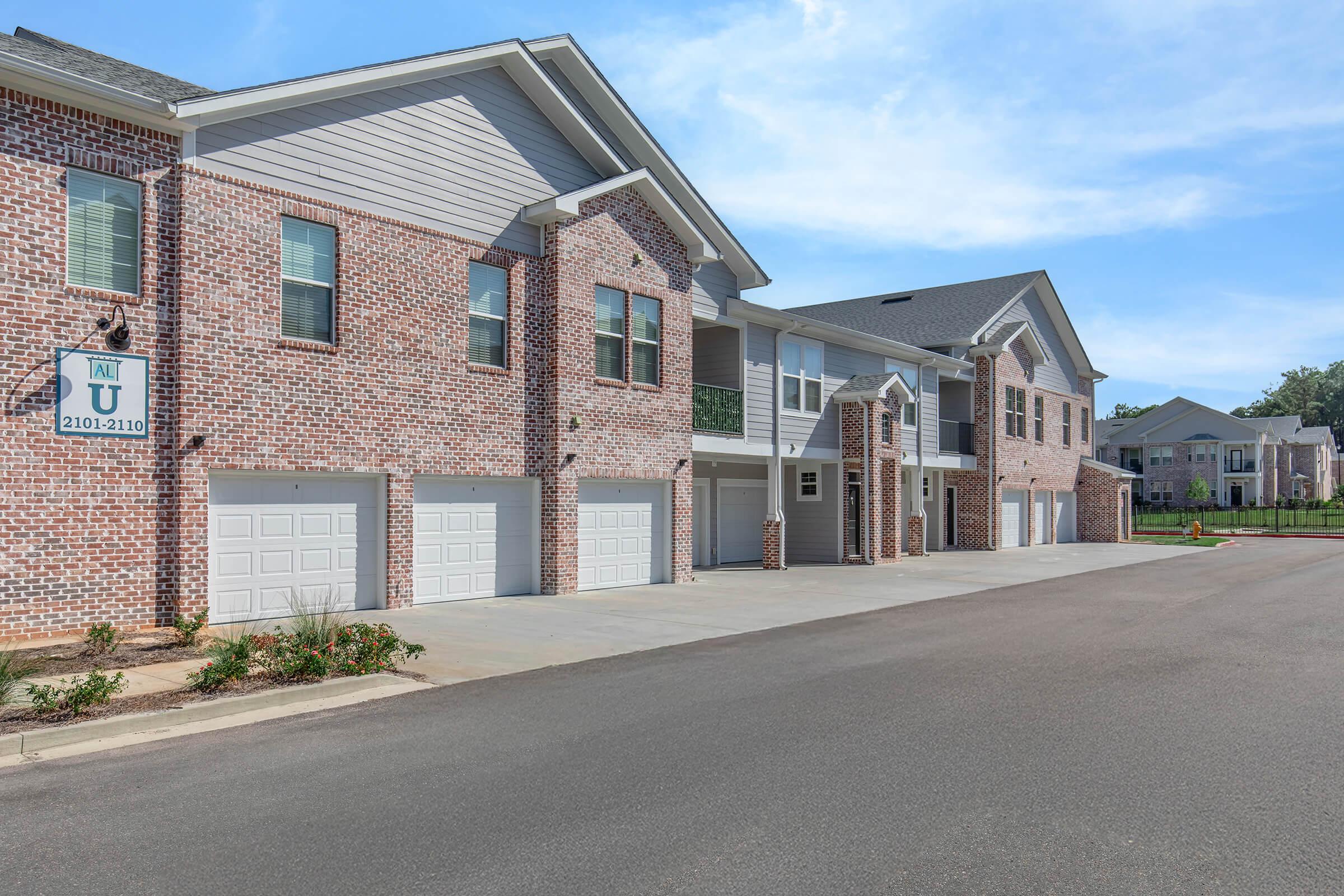 Brick apartment buildings with multiple garage doors lining a paved driveway. The sky is clear and blue, creating a bright atmosphere. There are freshly landscaped plants near the entrance, and additional apartment buildings are visible in the background.