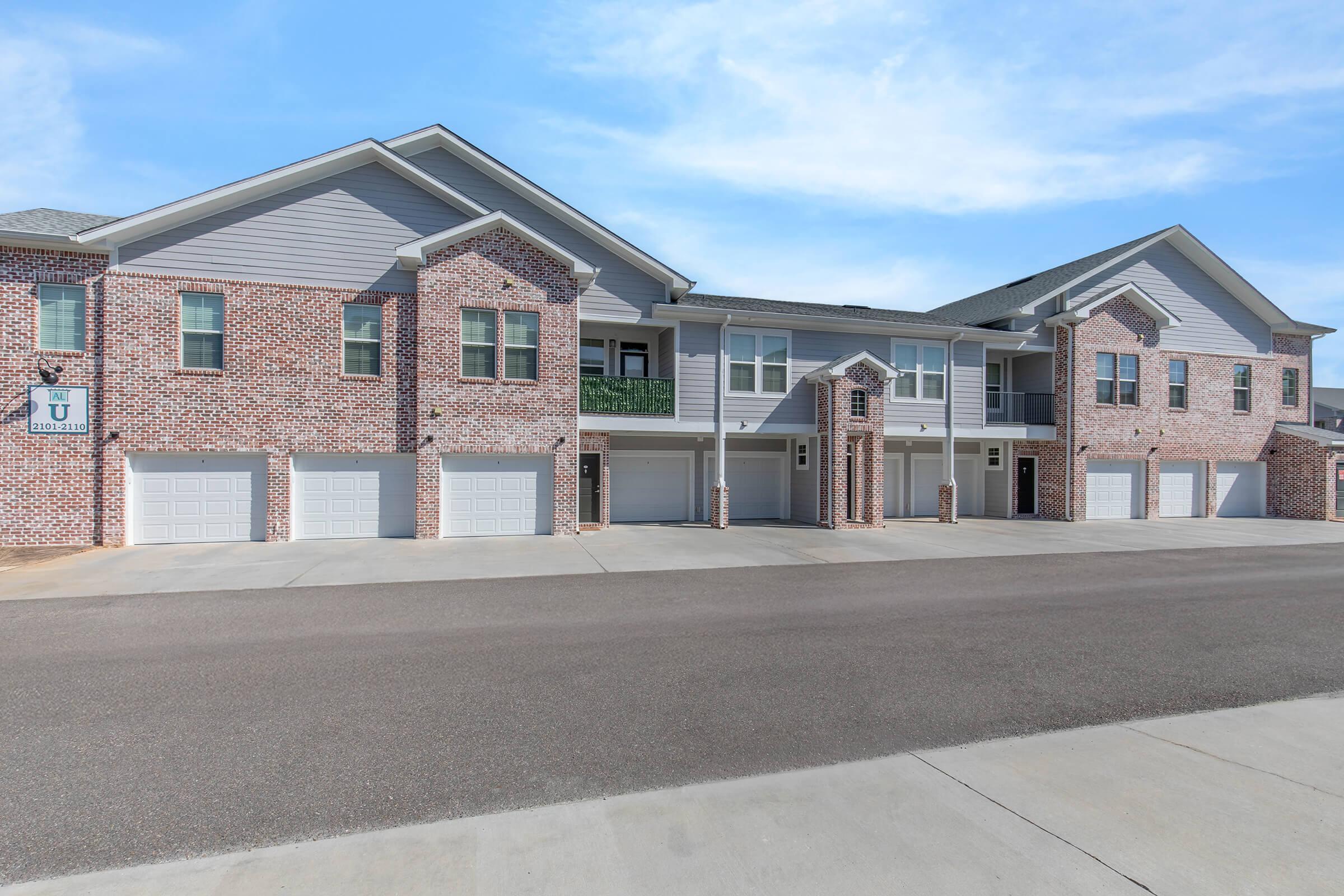 A modern brick apartment building with multiple units, each featuring a garage. The building has a light gray exterior with darker accents and a balcony on the second floor. The surrounding area is paved and clear, with a blue sky above.