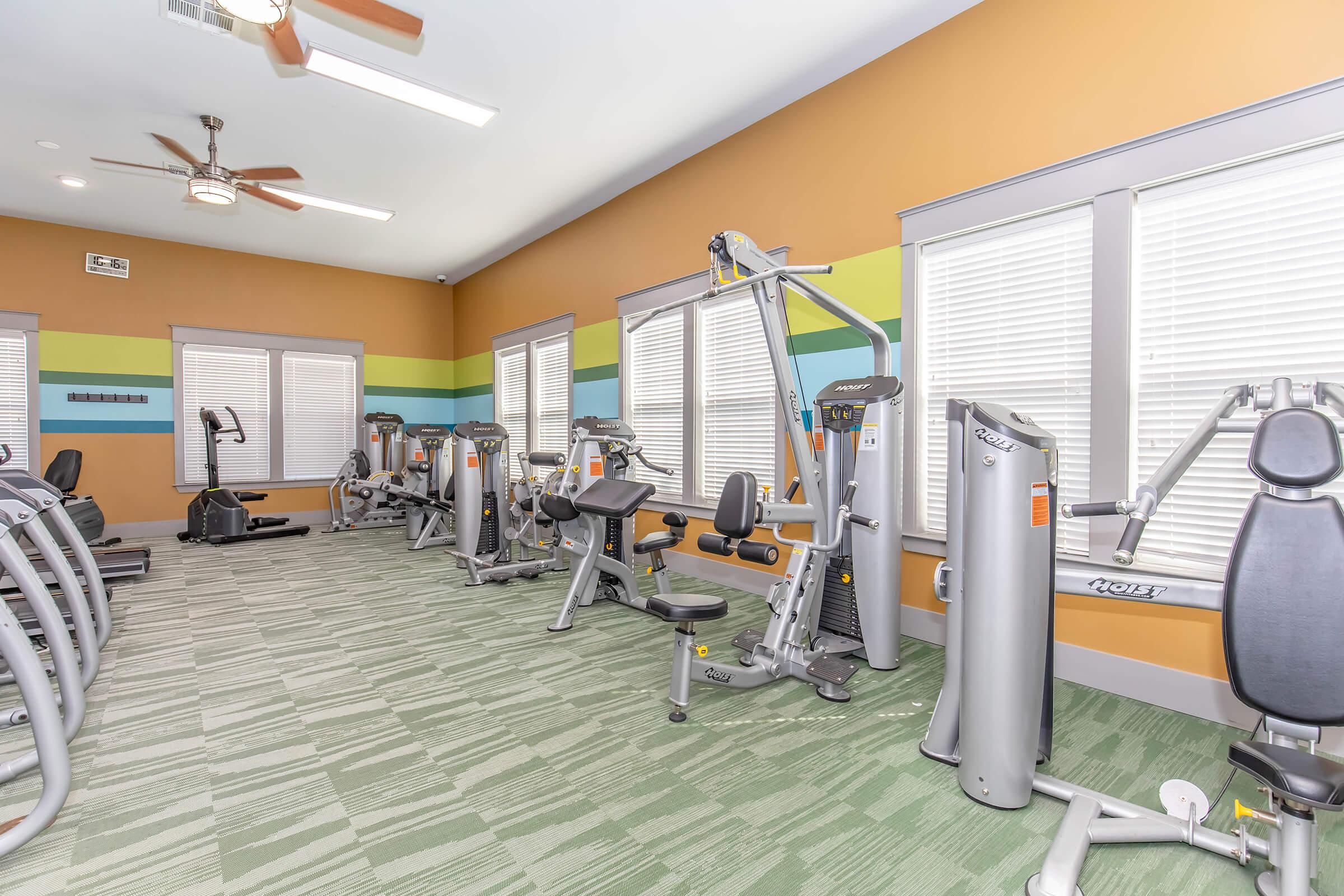 A modern gym interior featuring various exercise machines, including weightlifting and cardio equipment. The room is well-lit with multiple windows and decorated in bright colors. Green carpeting covers the floor, and there are ceiling fans for ventilation.