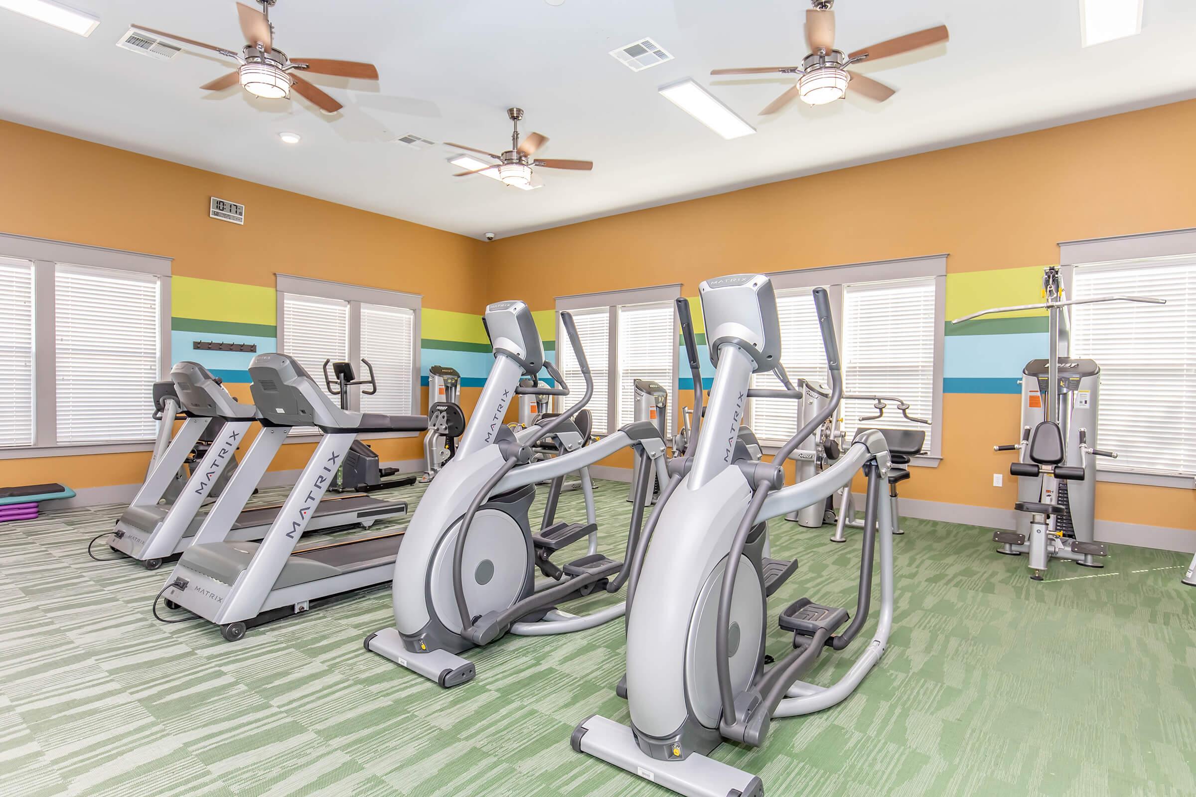 A well-lit gym space featuring modern exercise equipment, including ellipticals and a multi-gym machine. The walls are painted in warm colors with accent stripes, and there are ceiling fans for ventilation. Natural light filters through windows, creating an inviting atmosphere for workouts.