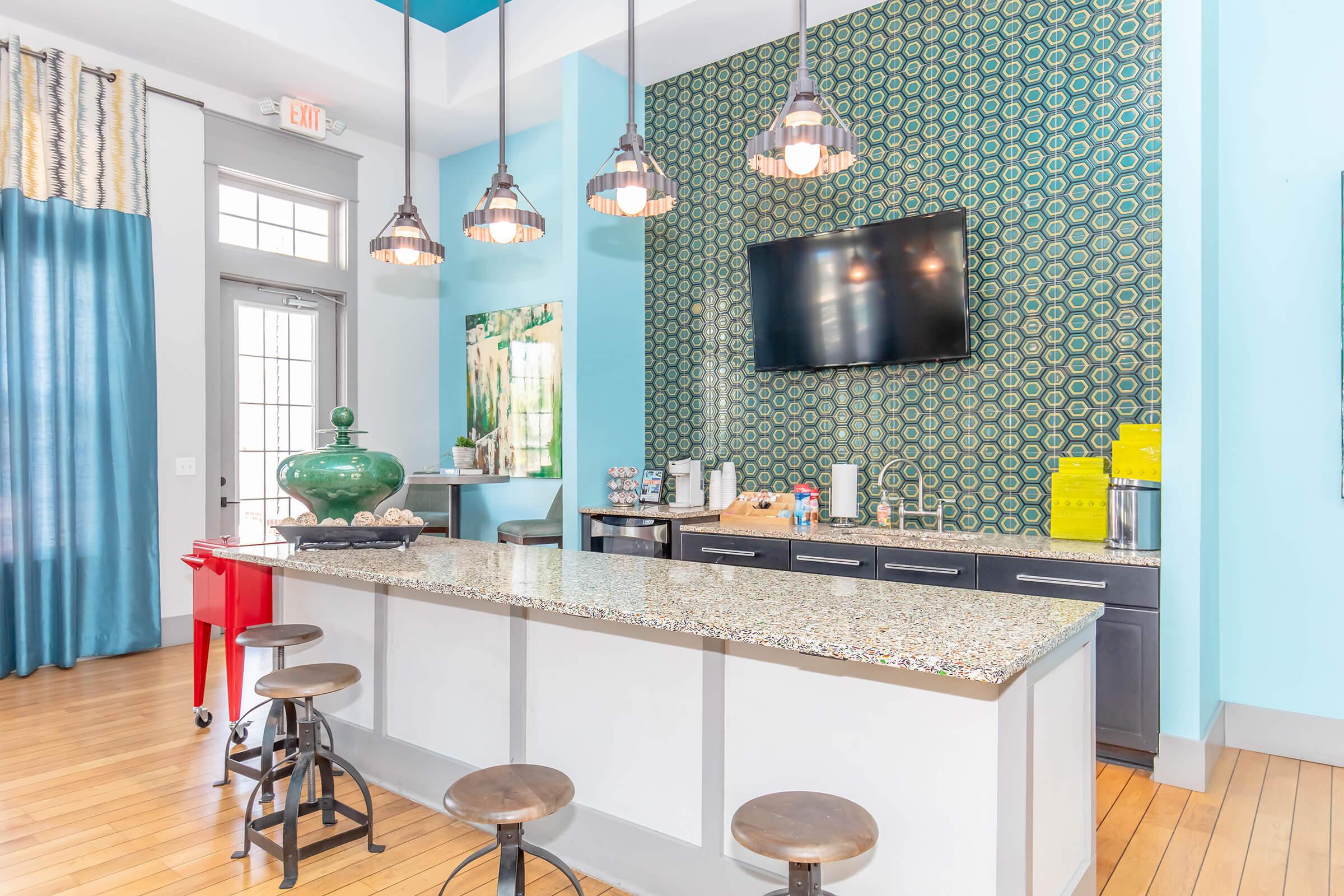 Modern kitchen area featuring a granite-topped island with stools, colorful patterned wall, a large television mounted above, and bright blue accents. The space is well-lit with pendant lights and has large windows, giving it a welcoming and airy atmosphere.