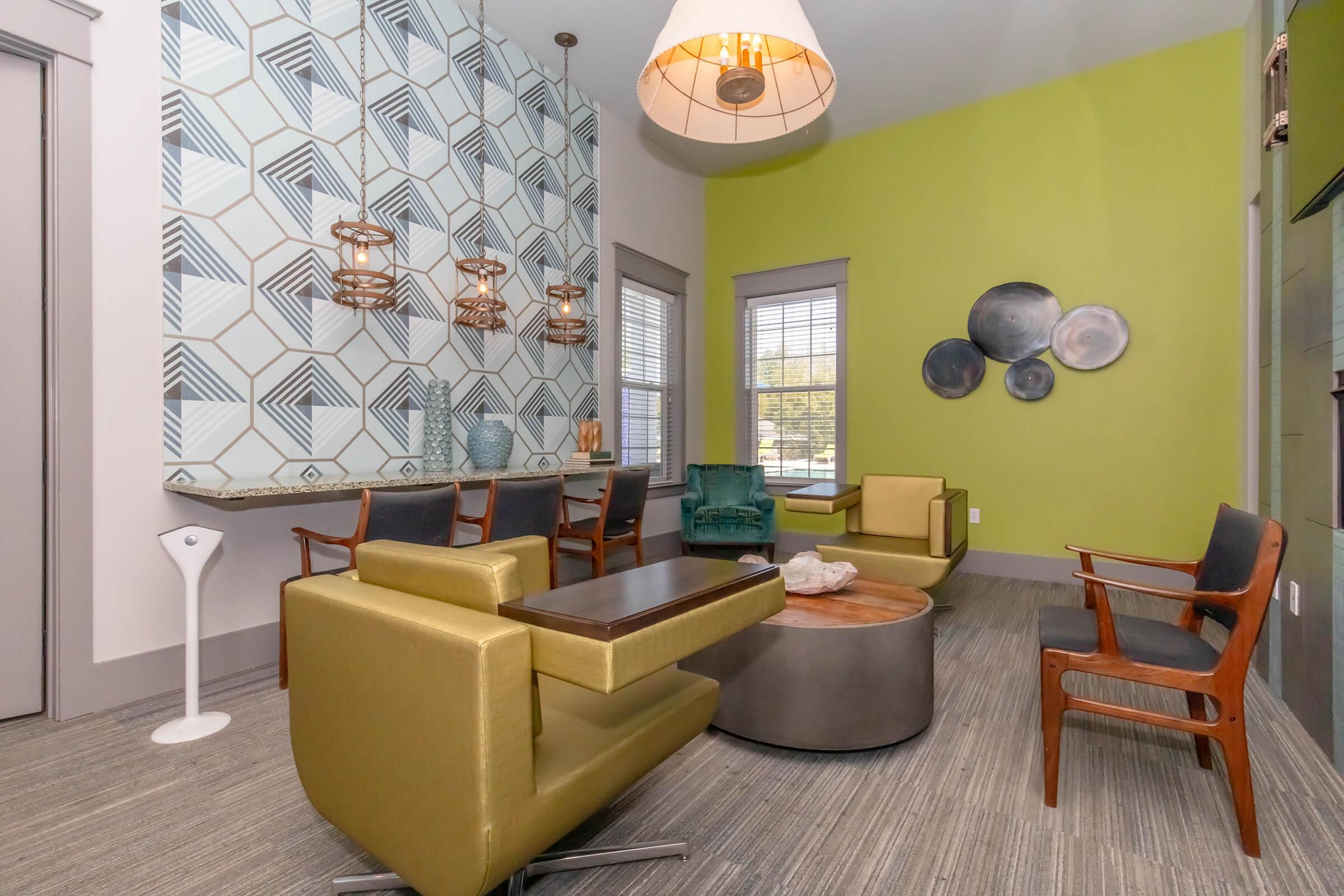 Modern lounge area featuring green and blue geometric wallpaper, stylish seating with a mix of green and gold chairs, a circular wooden coffee table, and decorative wall plates. Natural light streams in through large windows, creating a welcoming atmosphere.