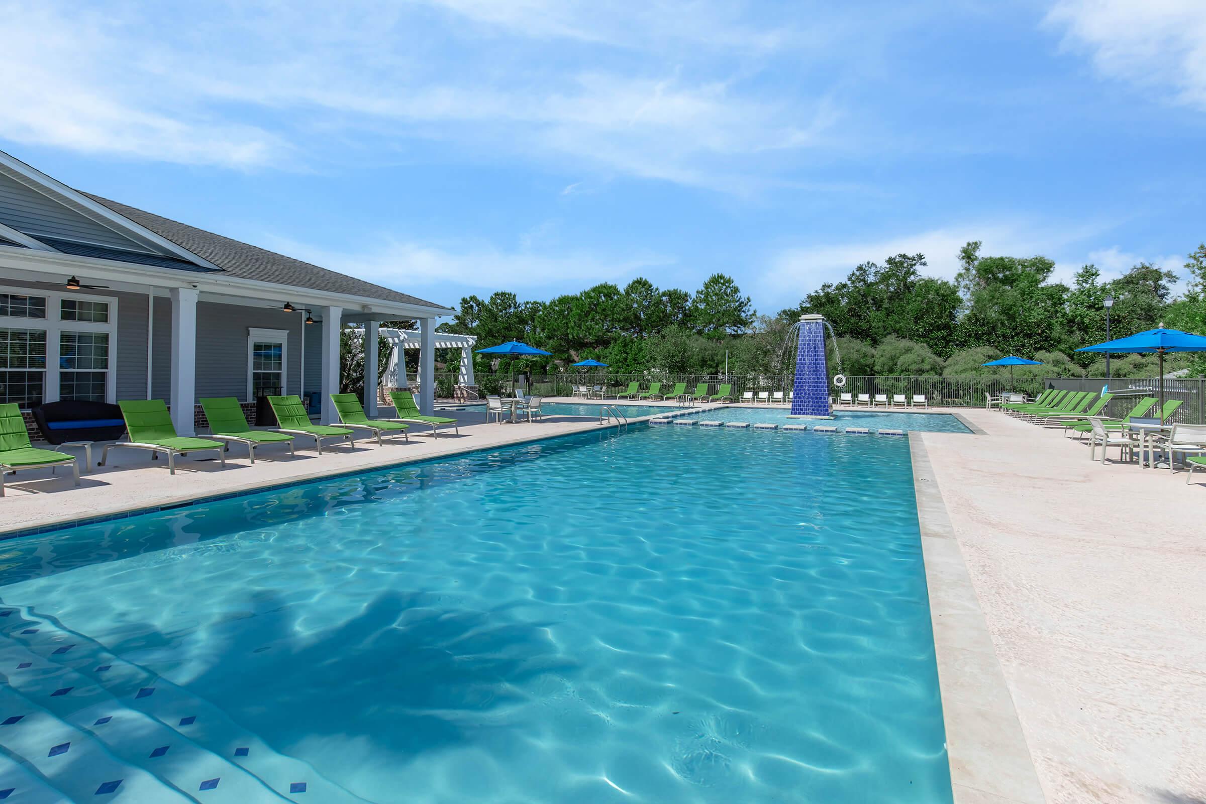 A serene outdoor swimming pool surrounded by lounge chairs and umbrellas. The pool features a blue water surface reflecting the sky, with a decorative water fountain at one end. Green trees and a clear sky create a relaxing atmosphere, ideal for leisure and recreation.