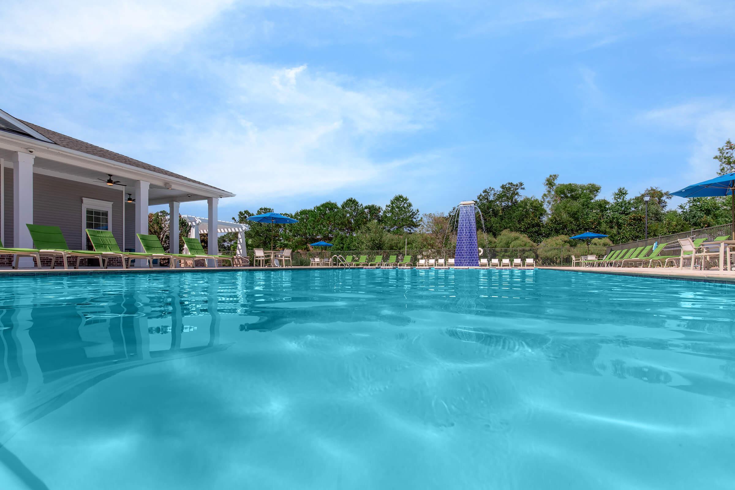 A clear blue swimming pool with lounge chairs lining the edge, surrounded by greenery and umbrellas. A fountain can be seen at one end of the pool, and the sky is bright with a few clouds, creating a serene and inviting atmosphere.