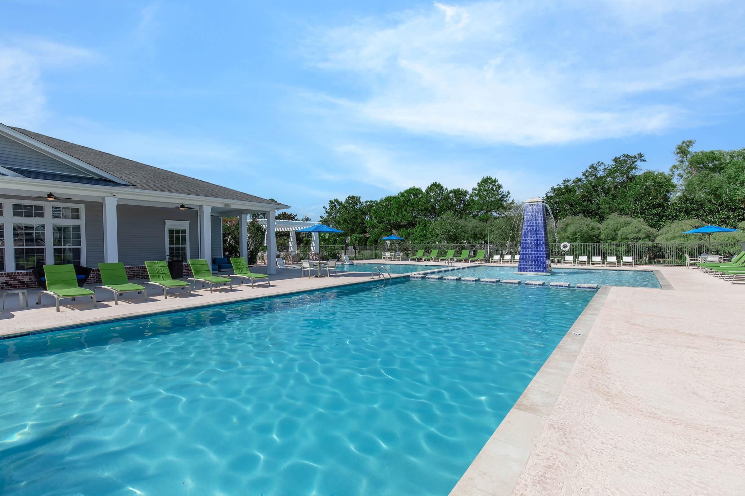 A bright, inviting swimming pool with crystal clear water, surrounded by green lounge chairs. In the background, there's a cascading water feature and a spacious patio area, complemented by blue umbrellas. Lush greenery can be seen beyond the pool area, under a clear blue sky.