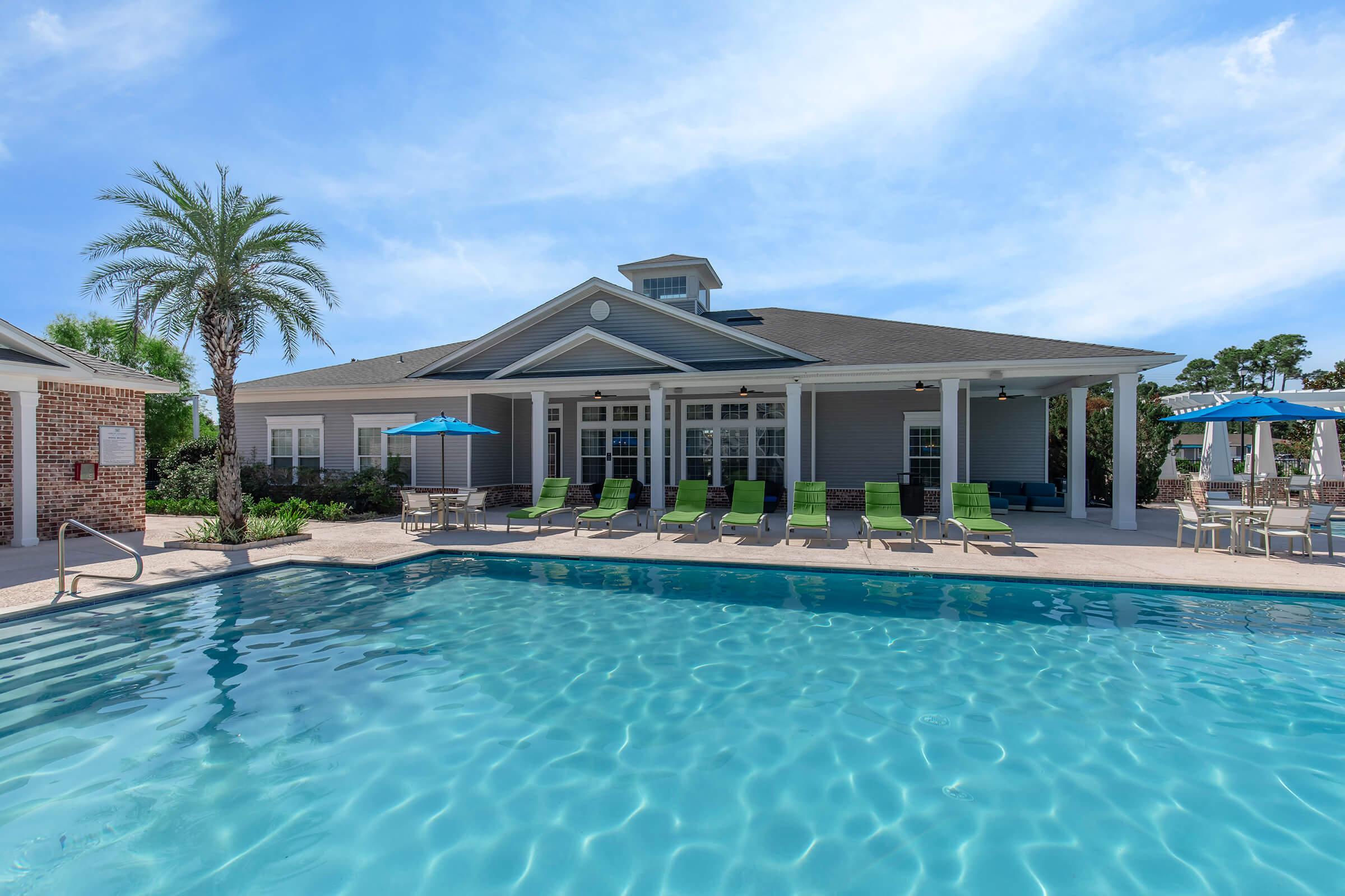 A clear blue swimming pool in front of a light gray building with large windows, surrounded by lounge chairs with bright green cushions. Palm trees and umbrellas add to the tropical ambiance, under a bright blue sky. The setting appears inviting and relaxed, ideal for leisure activities.