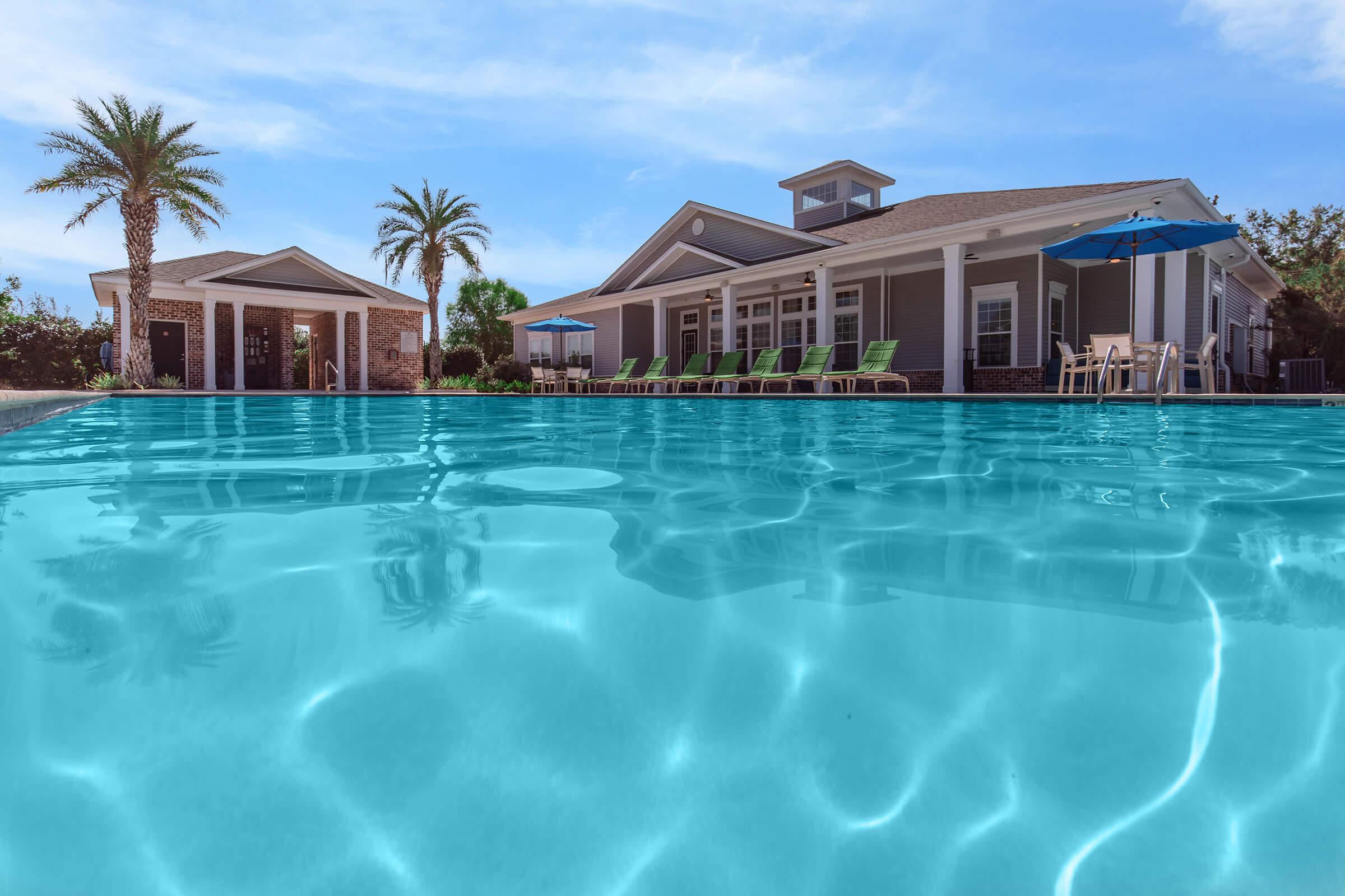 A serene swimming pool with turquoise water reflecting the sky, surrounded by palm trees. In the background, a stylish building with a porch and lounge chairs, along with blue umbrellas, creates a relaxing atmosphere ideal for leisure and enjoyment.