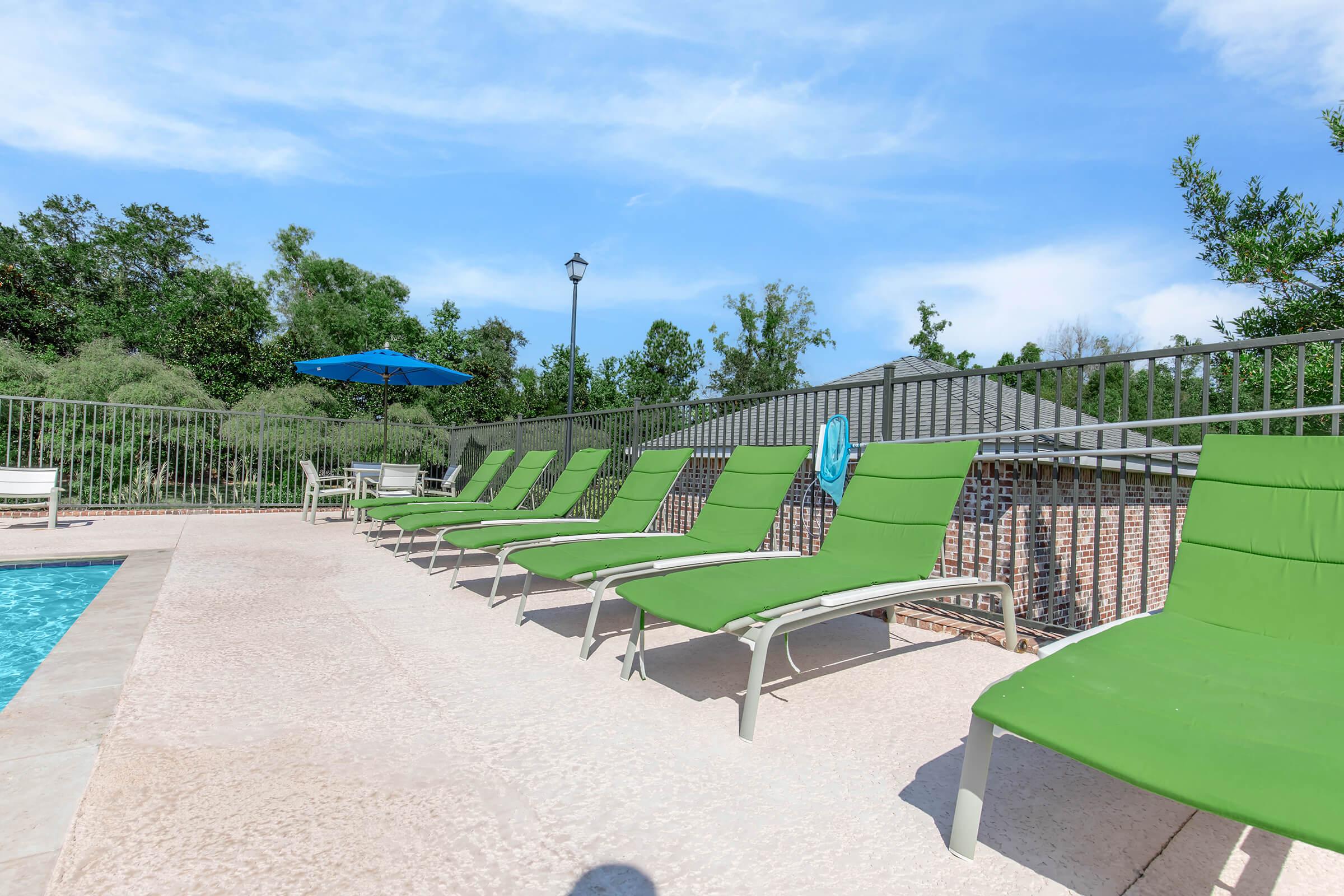 A poolside scene featuring several green lounge chairs arranged neatly along a pool. In the background, there are trees and a blue umbrella providing shade. The sky is clear with a few clouds, creating a bright and inviting atmosphere.