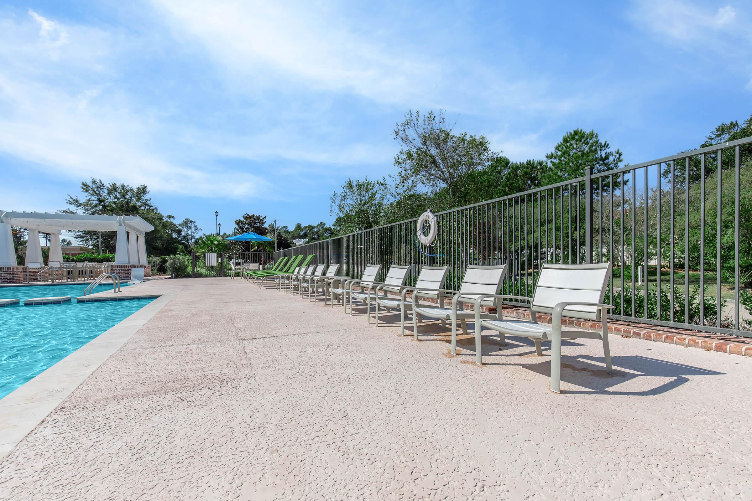 A clear blue swimming pool surrounded by lounge chairs on a patio. Green trees and a blue sky are visible in the background, creating a relaxing outdoor atmosphere. A lifebuoy is mounted on the fence near the pool area.