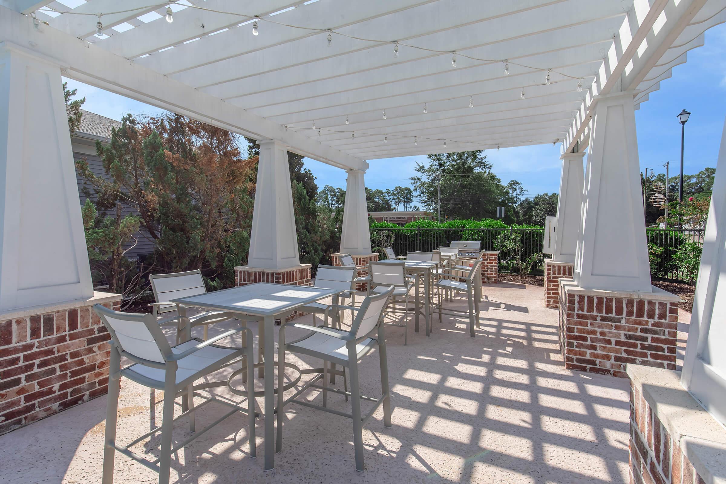 A shaded outdoor dining area with several square tables and chairs set on a light-colored surface, surrounded by green plants and trees. The structure features a white pergola with hanging lights, creating a cozy and inviting atmosphere. Sunlight casts shadows on the ground.