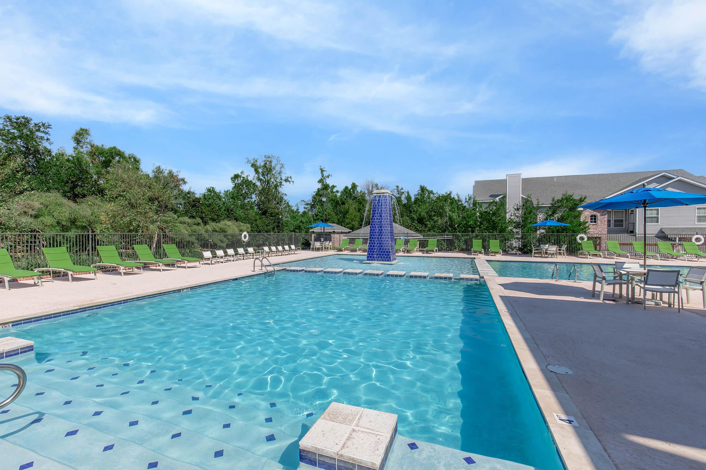 A clear blue swimming pool surrounded by green trees and lounge chairs. There is a water slide at one end of the pool, with umbrellas providing shade nearby. The area is sunny and inviting, perfect for relaxation and swimming.
