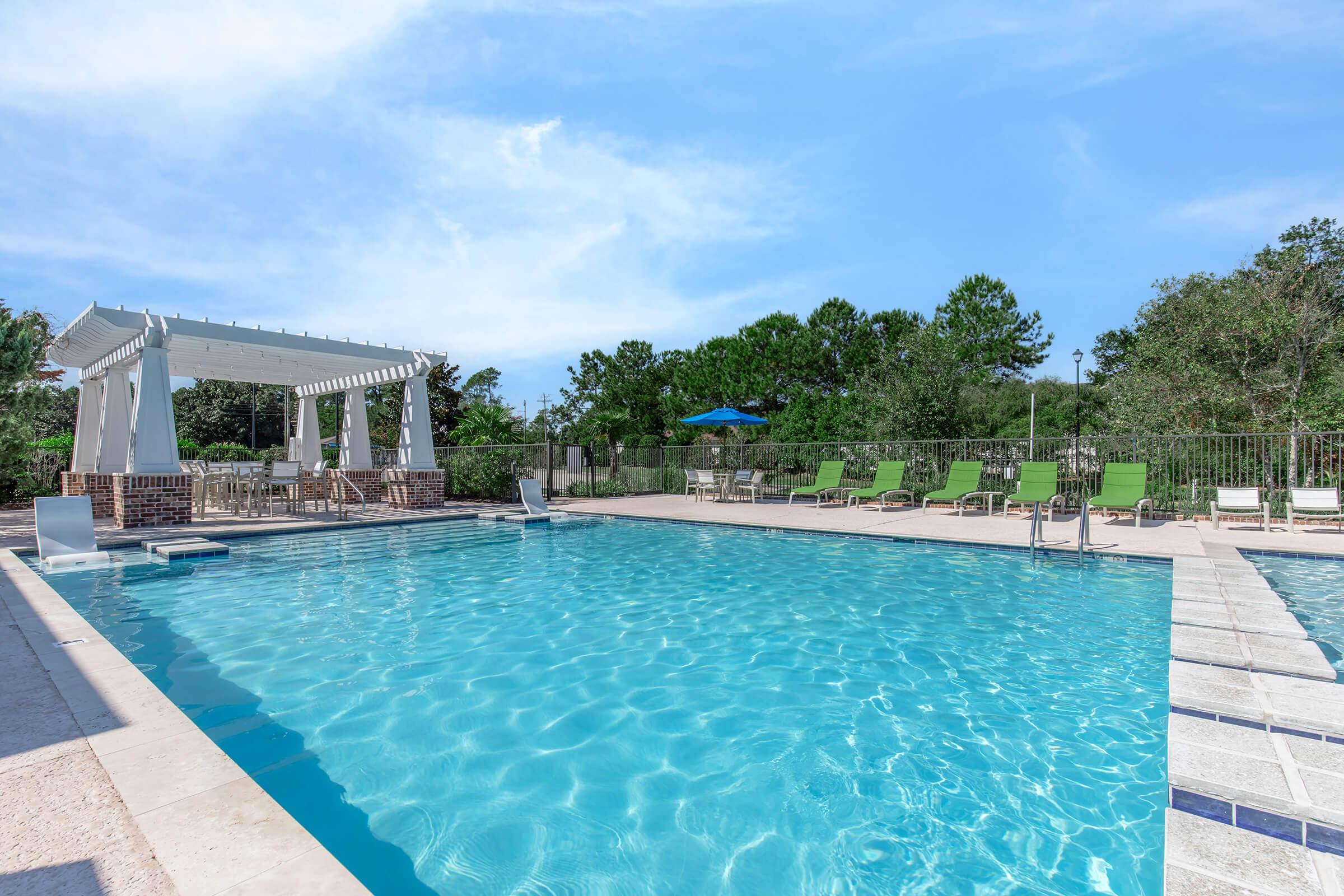 A clear blue swimming pool surrounded by green lounge chairs and trees. There’s a shaded area with a white gazebo and a blue umbrella in the background, under a bright blue sky. The pool area is well-maintained and inviting, perfect for relaxation and recreation.
