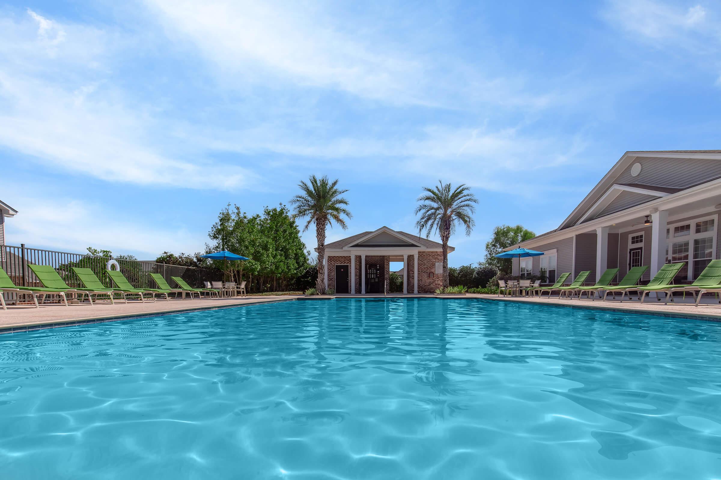 A clear blue swimming pool with sun loungers arranged around it, surrounded by palm trees. In the background, there is a shaded gazebo and a building with a porch. The sky is bright and partially cloudy, creating a relaxed outdoor atmosphere.