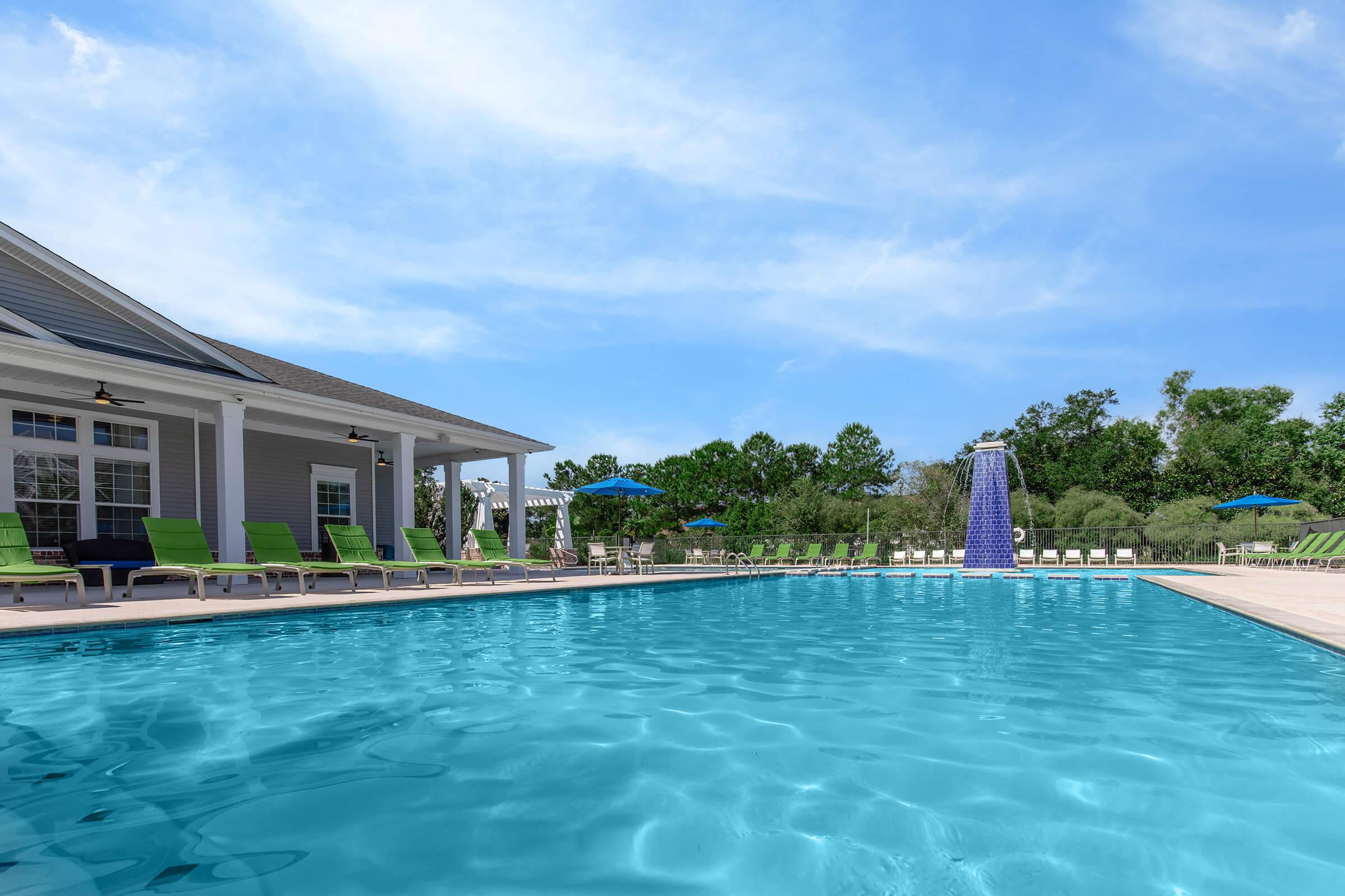 A clear blue swimming pool with lounge chairs and green umbrellas surrounding it. In the background, there is a modern building with large windows. A waterfall feature is visible at one end of the pool, and lush greenery is seen in the distance under a clear blue sky.