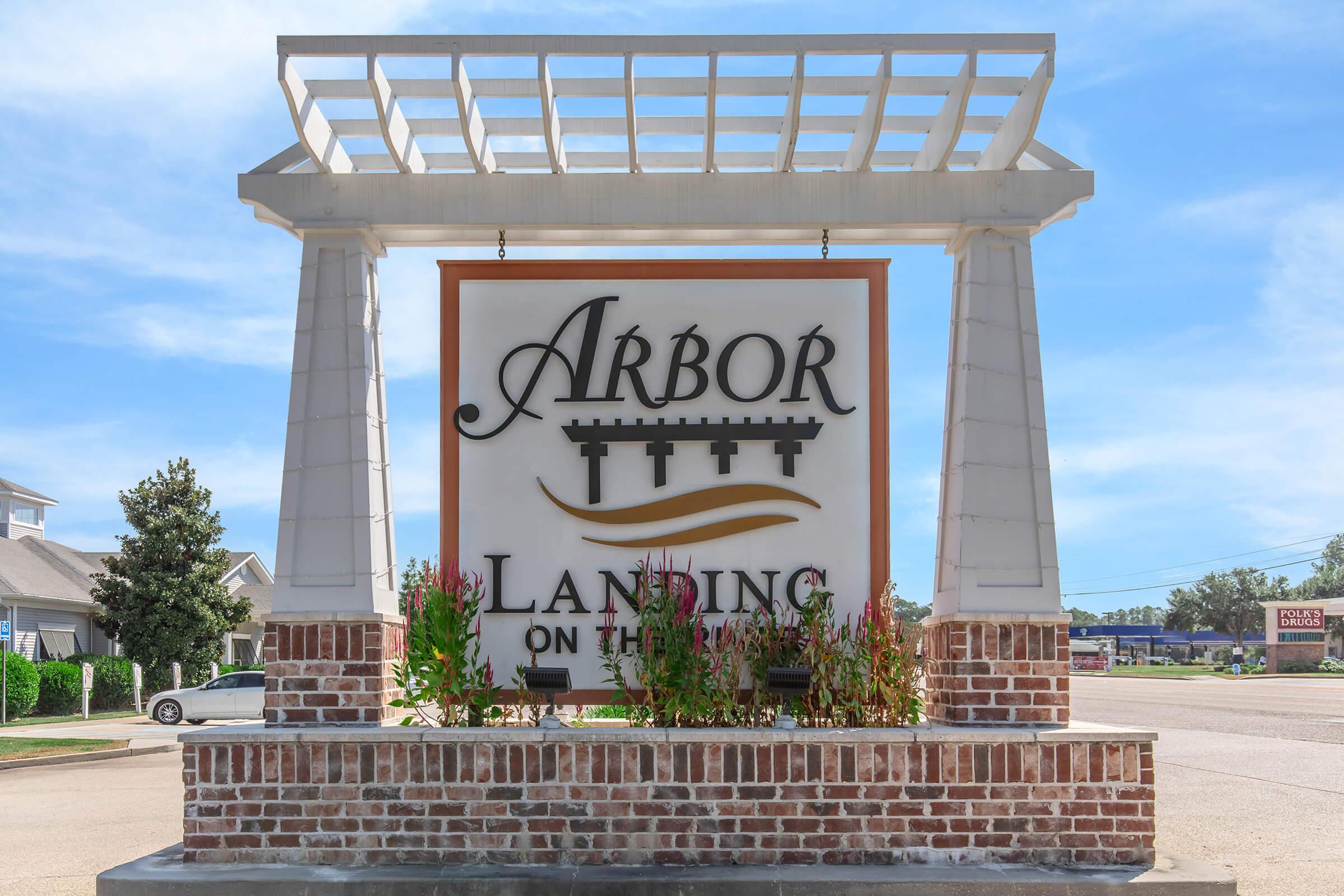 Sign for Arbor Landing on the River, featuring elegant lettering and decorative elements. Surrounded by brickwork and landscaped plants, the sign is set against a clear blue sky, indicating a welcoming entrance to the property.