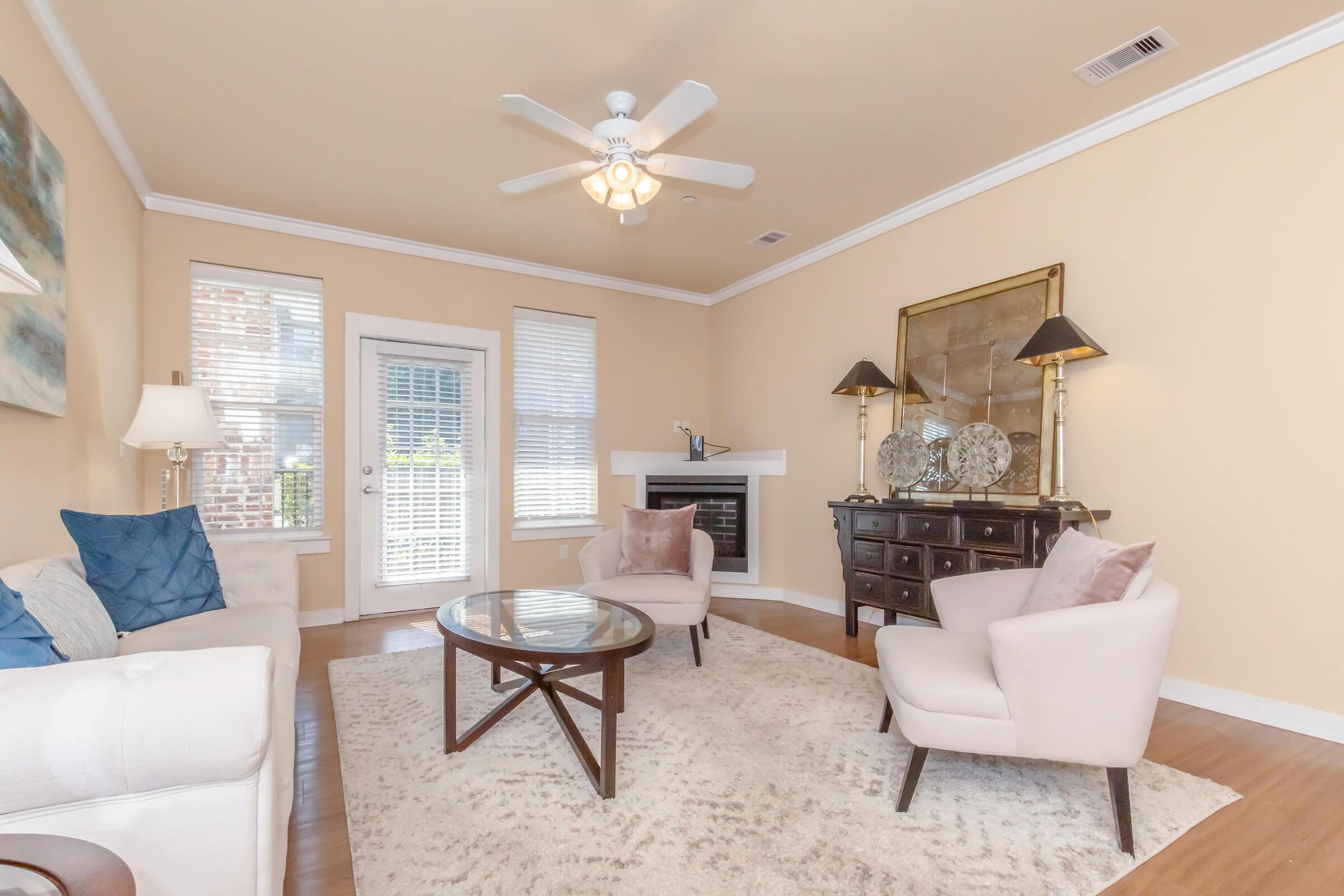 A cozy living room featuring a light-colored sofa with blue and white pillows, two beige armchairs, a round glass coffee table, and a large area rug. A ceiling fan hangs above, and a decorative fireplace is visible. Large windows let in natural light, and an elegant wall decor piece adds charm.