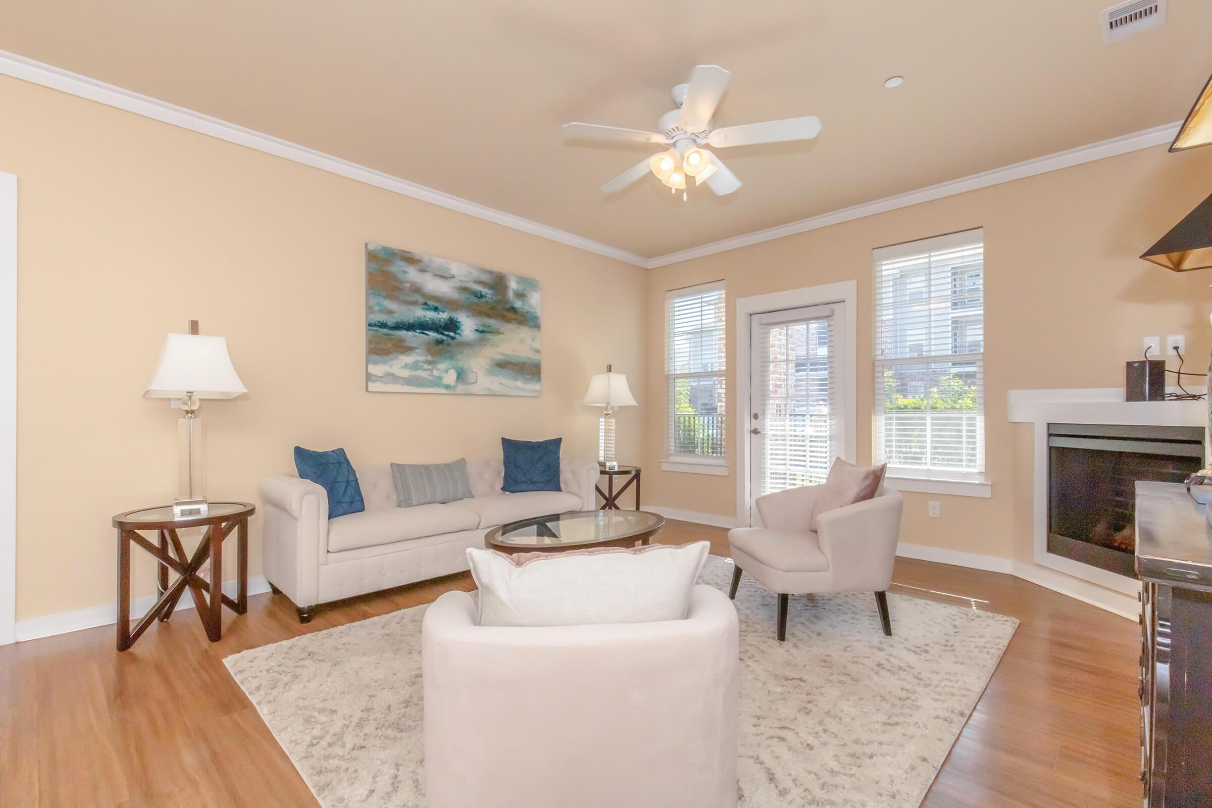 A cozy living room featuring a light beige color scheme, a white sofa with blue accent pillows, a round glass coffee table, and an armchair. There is a ceiling fan above and large windows with white blinds letting in natural light. A decorative painting hangs on the wall, and a fireplace adds warmth to the space.