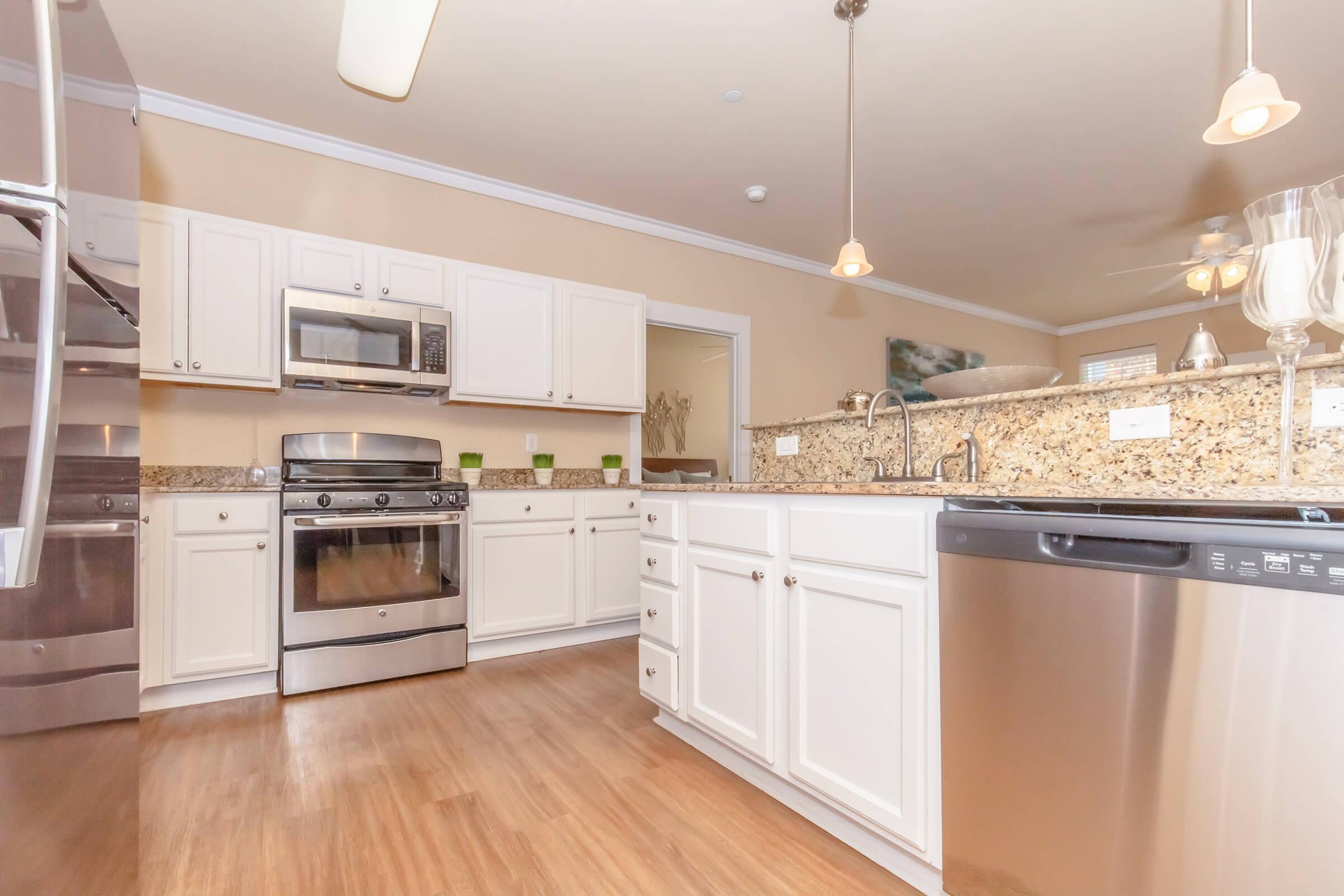 Modern kitchen featuring stainless steel appliances, including a microwave, oven, and refrigerator. The space has white cabinetry, granite countertops, and a dishwasher, with warm hardwood flooring and soft lighting from pendant fixtures.