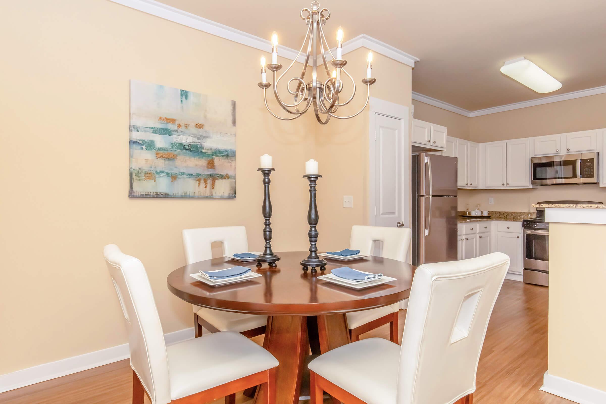 A modern dining area featuring a round wooden table with four white chairs, set with light blue plates. Above hangs an elegant chandelier, and a colorful abstract painting is mounted on the wall. The kitchen area is partially visible in the background, with white cabinets and stainless steel appliances.