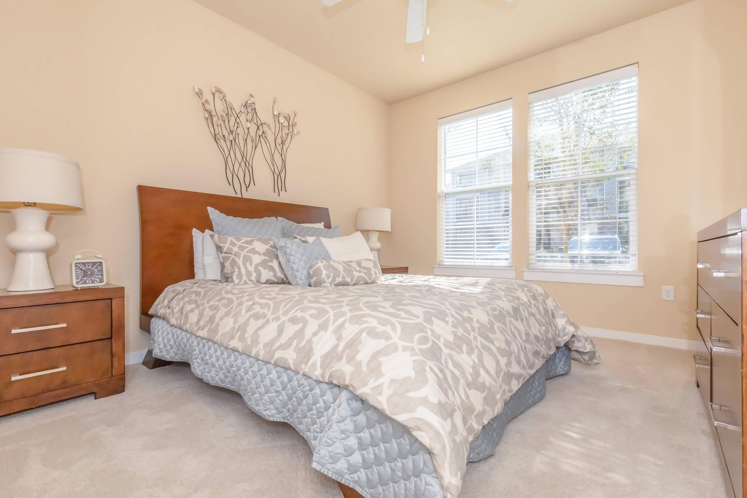 A cozy bedroom featuring a king-size bed with a patterned gray and white comforter. There are two bedside lamps, a wooden dresser, and a decorative wall piece. Natural light streams through two large windows, illuminating the soft, neutral-toned walls and light carpet.