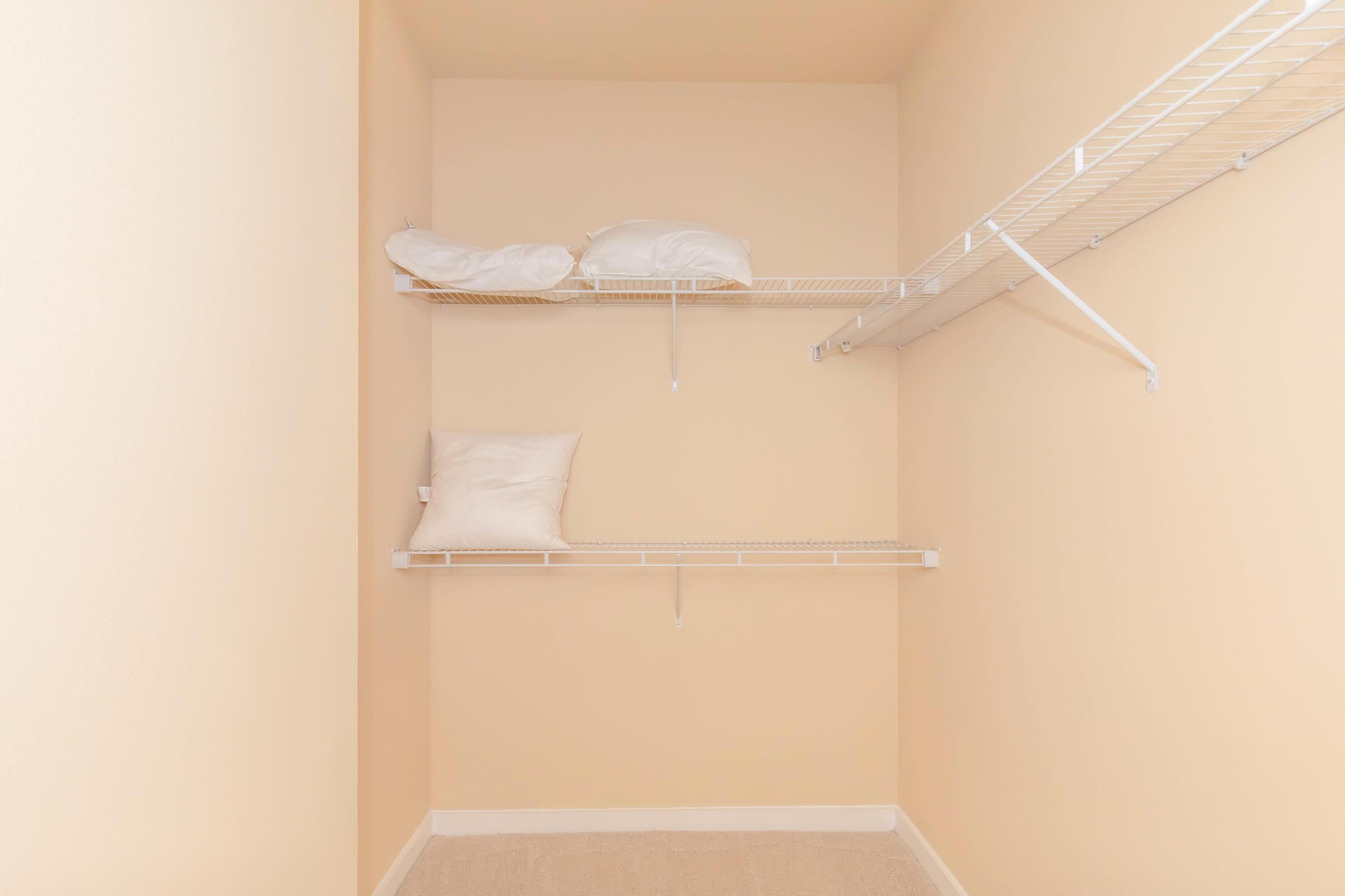 A neatly organized closet featuring light beige walls. It has two wire shelves, one with a couple of white pillows resting on it, and the other shelf empty. The floor is carpeted in a neutral tone, creating a simple and clean appearance.