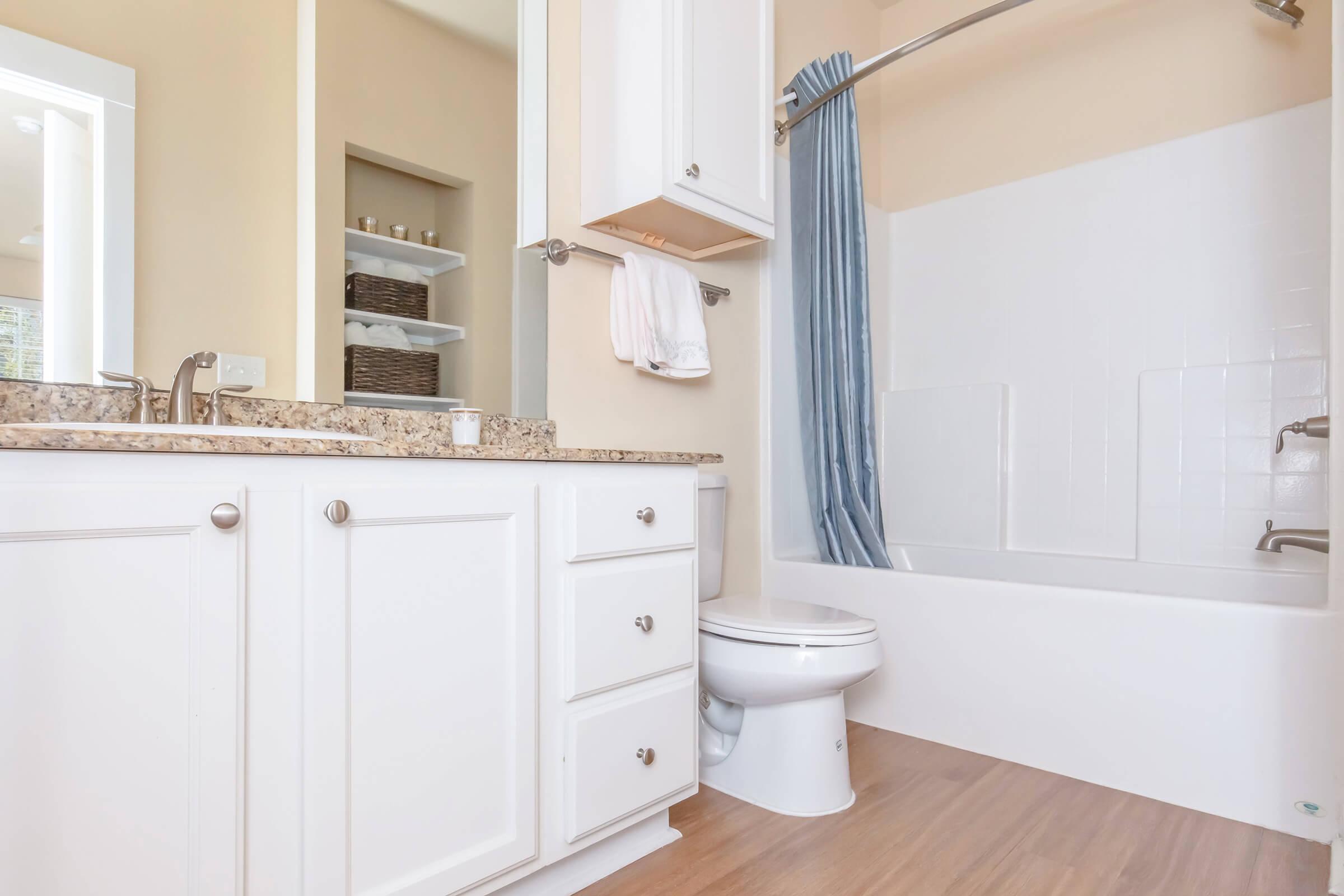 A clean bathroom featuring a white bathtub with a blue curtain, a modern toilet, and a granite countertop with storage. Shelves in the background hold woven baskets. Natural light enters through a nearby window, highlighting the neutral color scheme.