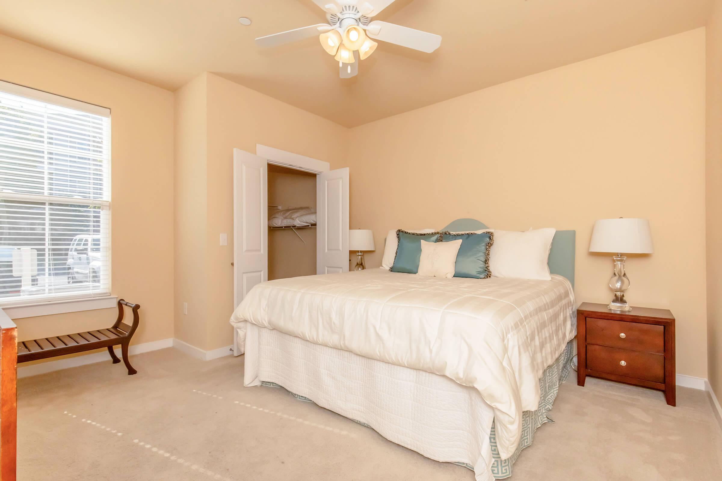Bright and airy bedroom featuring a large bed with decorative pillows, a bedside table with a lamp, and a chair by the window. The walls are a warm beige, and a ceiling fan provides airflow. A closet is visible, offering additional storage space, and natural light streams in through the window.