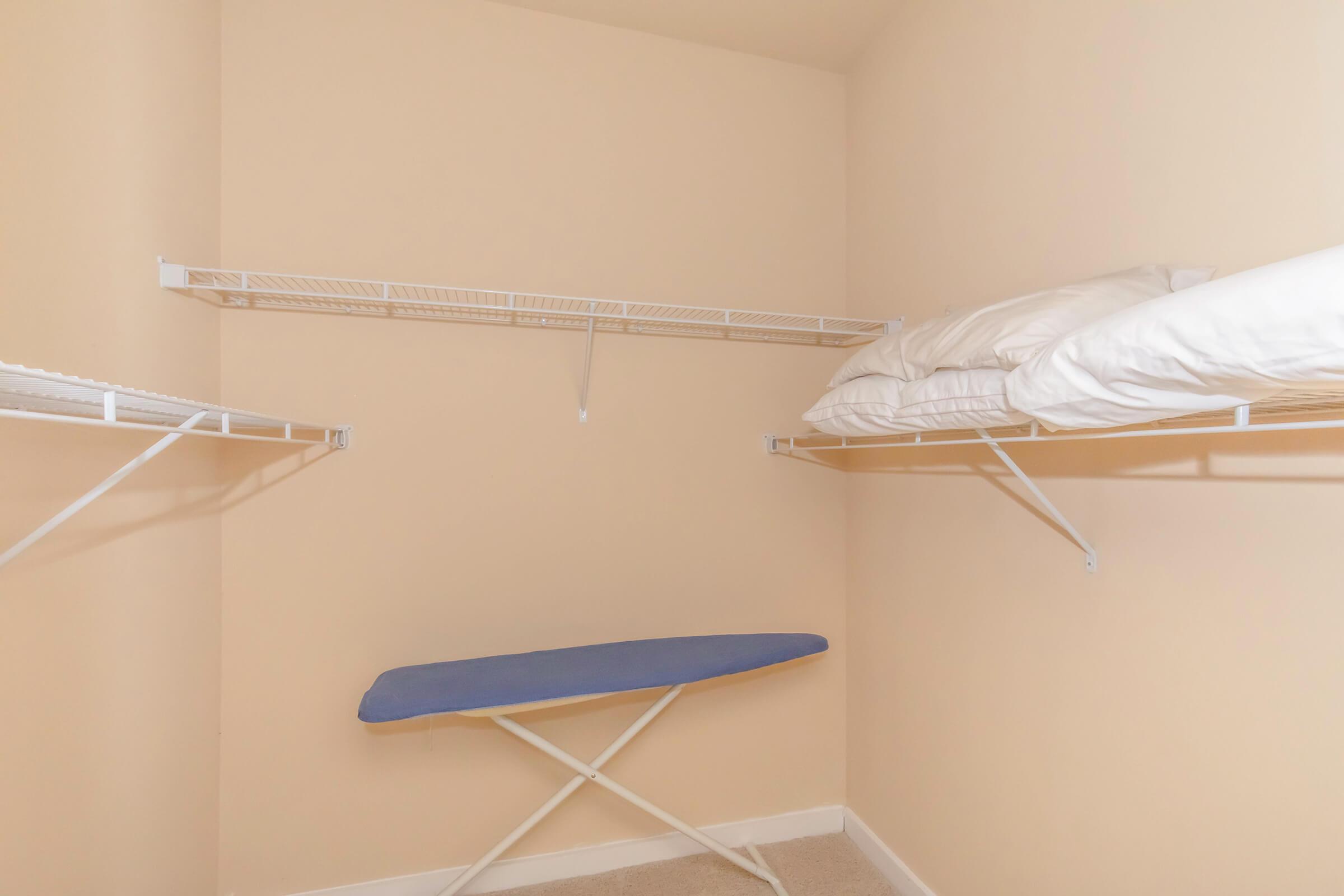 A small closet with light beige walls. It features two wire shelves on the top for storage, and an ironing board with a blue cover positioned on the floor. Two white pillows are neatly stacked on one of the shelves. The flooring is carpeted in a neutral color.